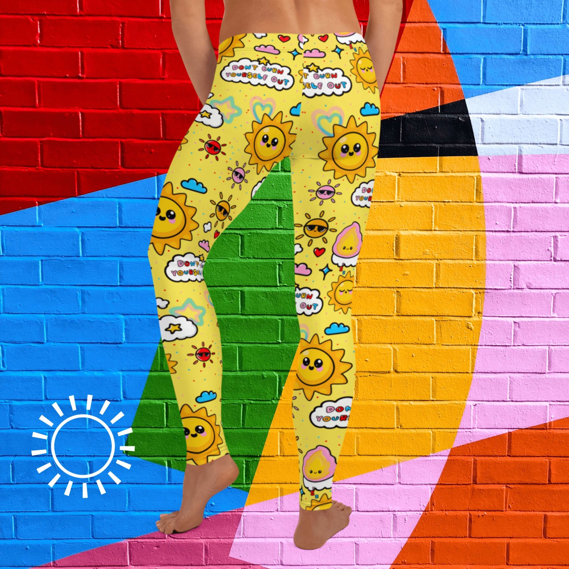 The Don't Burn Yourself Out Leggings on a model over a rainbow brick wall background with a white sunshine outline in the bottom left corner. The model is facing away with their left leg bent and both hands resting by their side. The print is yellow with various kawaii face sunshines, sparkles, hearts, pink flames and pink, blue and white clouds with don't burn yourself out inside in rainbow. Design inspired by the self care movement.
