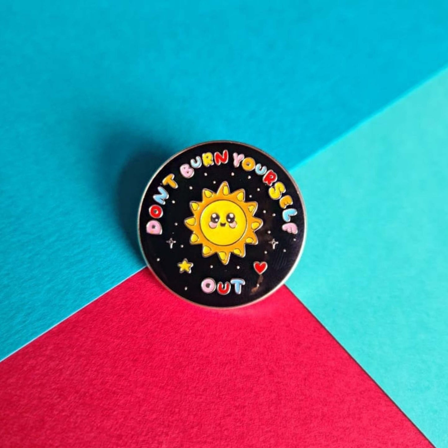 The Don't Burn Yourself Out Sunshine Enamel Pin on a red and blue background. The circle shaped pin badge is a black base with a yellow smiling sunshine in the centre with rainbow text surrounding it reading 'don't burn yourself out' along with a red heart, yellow star and silver sparkles. The design was created as a gentle reminder to practise self care.