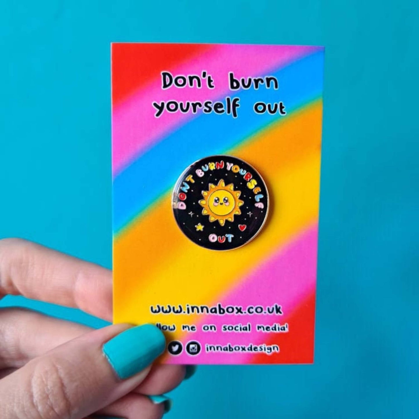 The Don't Burn Yourself Out Sunshine Enamel Pin on rainbow backing card with black top text reading 'don't burn yourself out' and bottom black text of the innabox website and social media handles, being held over a blue background. Circle shaped pin badge is a black base with a yellow smiling sunshine in the centre with rainbow text surrounding it reading 'don't burn yourself out' along with a red heart, yellow star and silver sparkles. The design was created as a gentle reminder to practise self care.