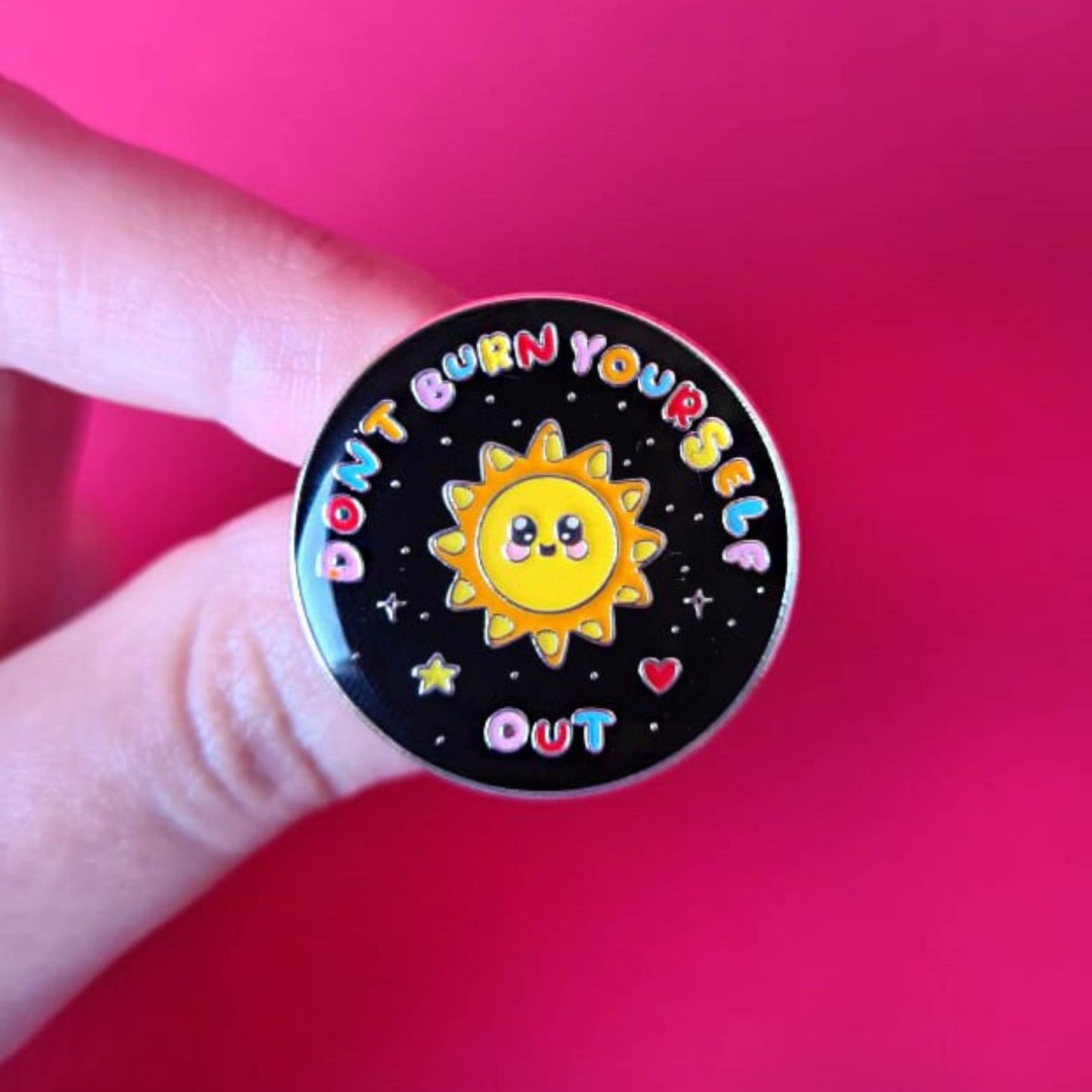 The Don't Burn Yourself Out Sunshine Enamel Pin being held over a red background. The circle shaped pin badge is a black base with a yellow smiling sunshine in the centre with rainbow text surrounding it reading 'don't burn yourself out' along with a red heart, yellow star and silver sparkles. The design was created as a gentle reminder to practise self care.