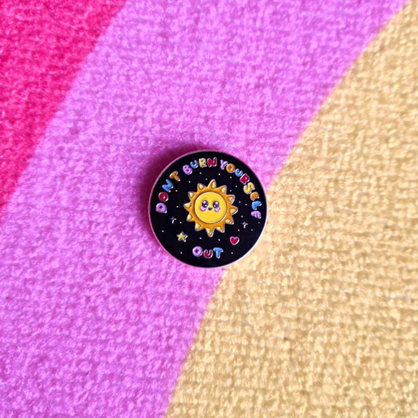 The Don't Burn Yourself Out Sunshine Enamel Pin on a rainbow fluffy rug. The circle shaped pin badge is a black base with a yellow smiling sunshine in the centre with rainbow text surrounding it reading 'don't burn yourself out' along with a red heart, yellow star and silver sparkles. The design was created as a gentle reminder to practise self care.