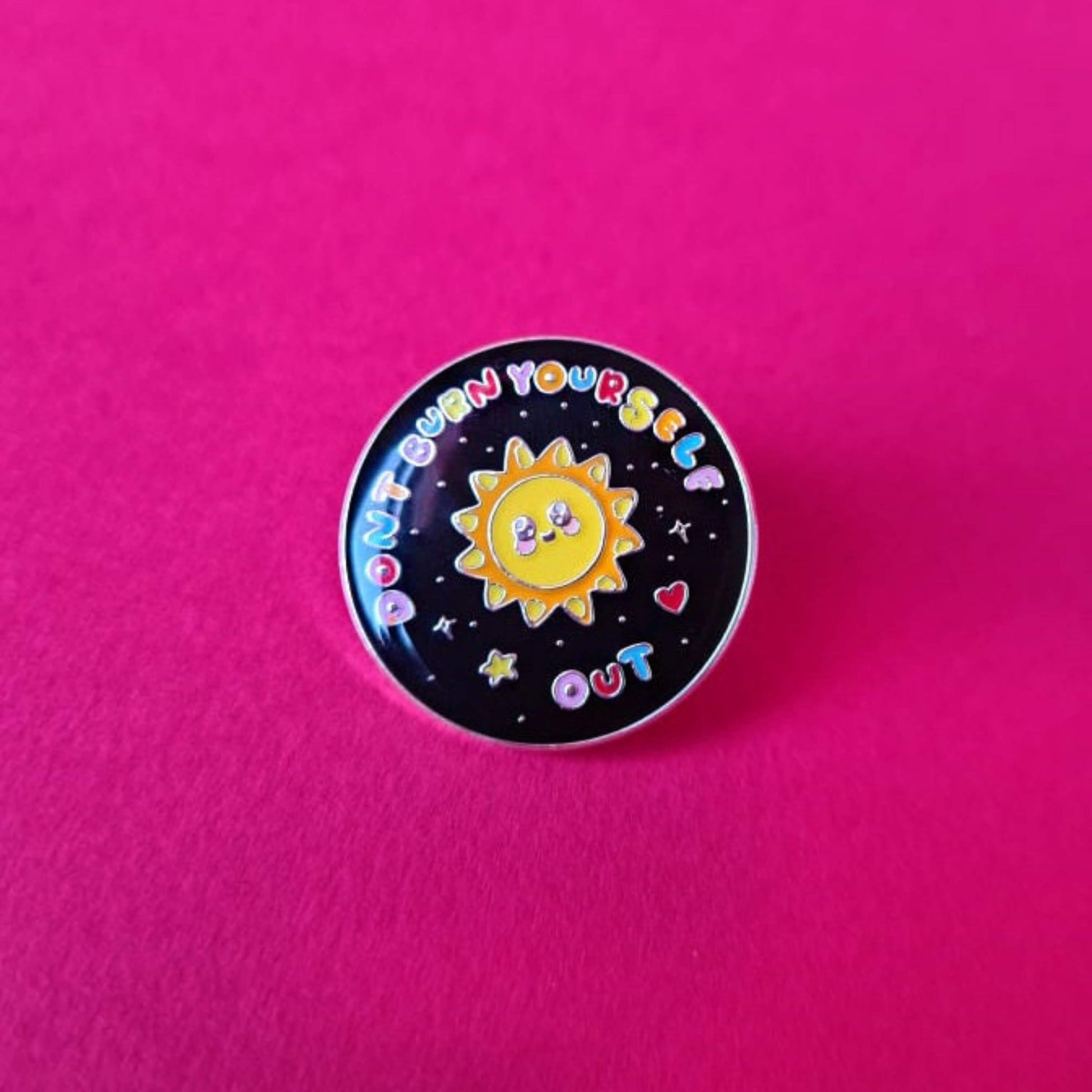 The Don't Burn Yourself Out Sunshine Enamel Pin being on a red background. The circle shaped pin badge is a black base with a yellow smiling sunshine in the centre with rainbow text surrounding it reading 'don't burn yourself out' along with a red heart, yellow star and silver sparkles. The design was created as a gentle reminder to practise self care.