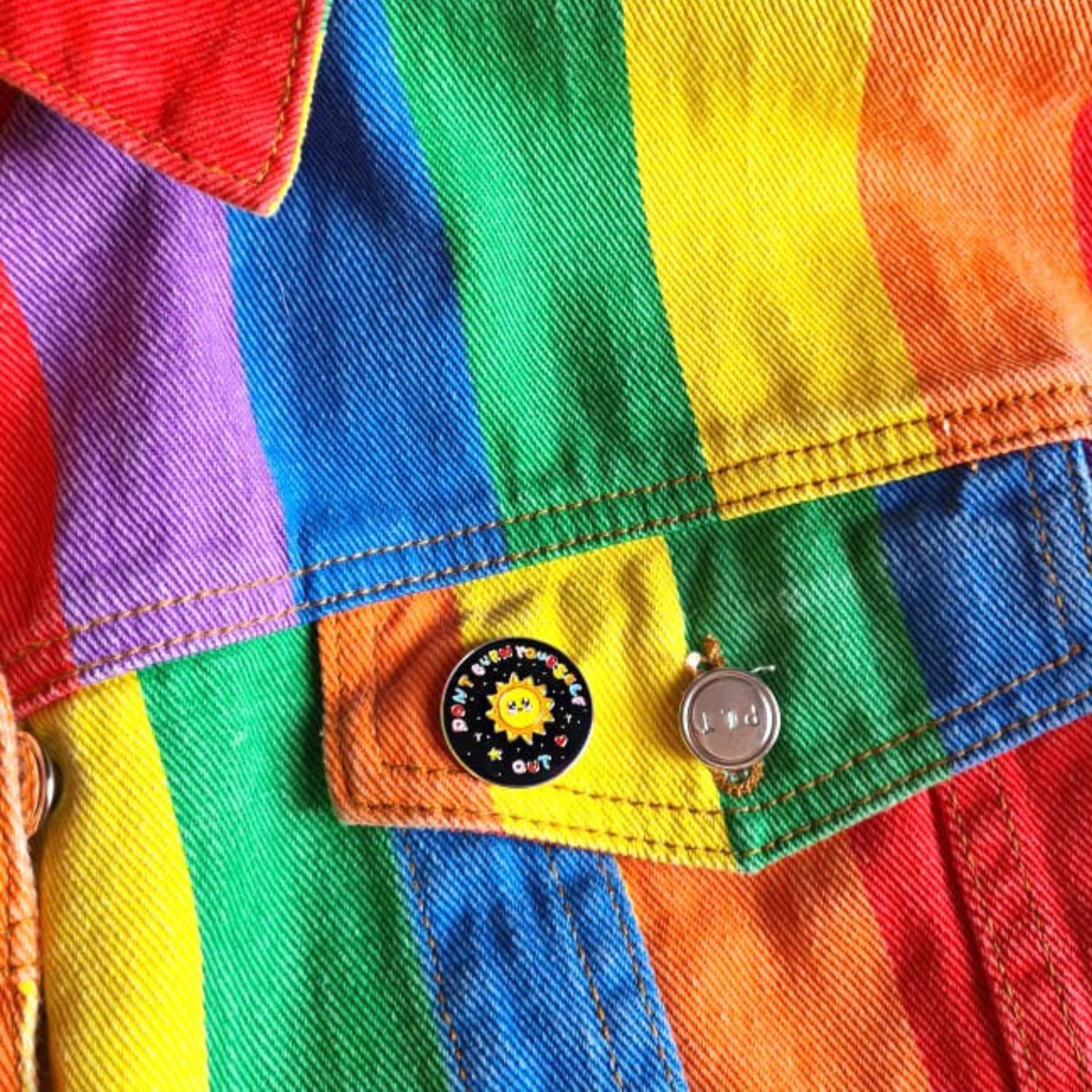 The Don't Burn Yourself Out Sunshine Enamel Pin pinned on a pocket of a rainbow denim jacket. The circle shaped pin badge is a black base with a yellow smiling sunshine in the centre with rainbow text surrounding it reading 'don't burn yourself out' along with a red heart, yellow star and silver sparkles. The design was created as a gentle reminder to practise self care.