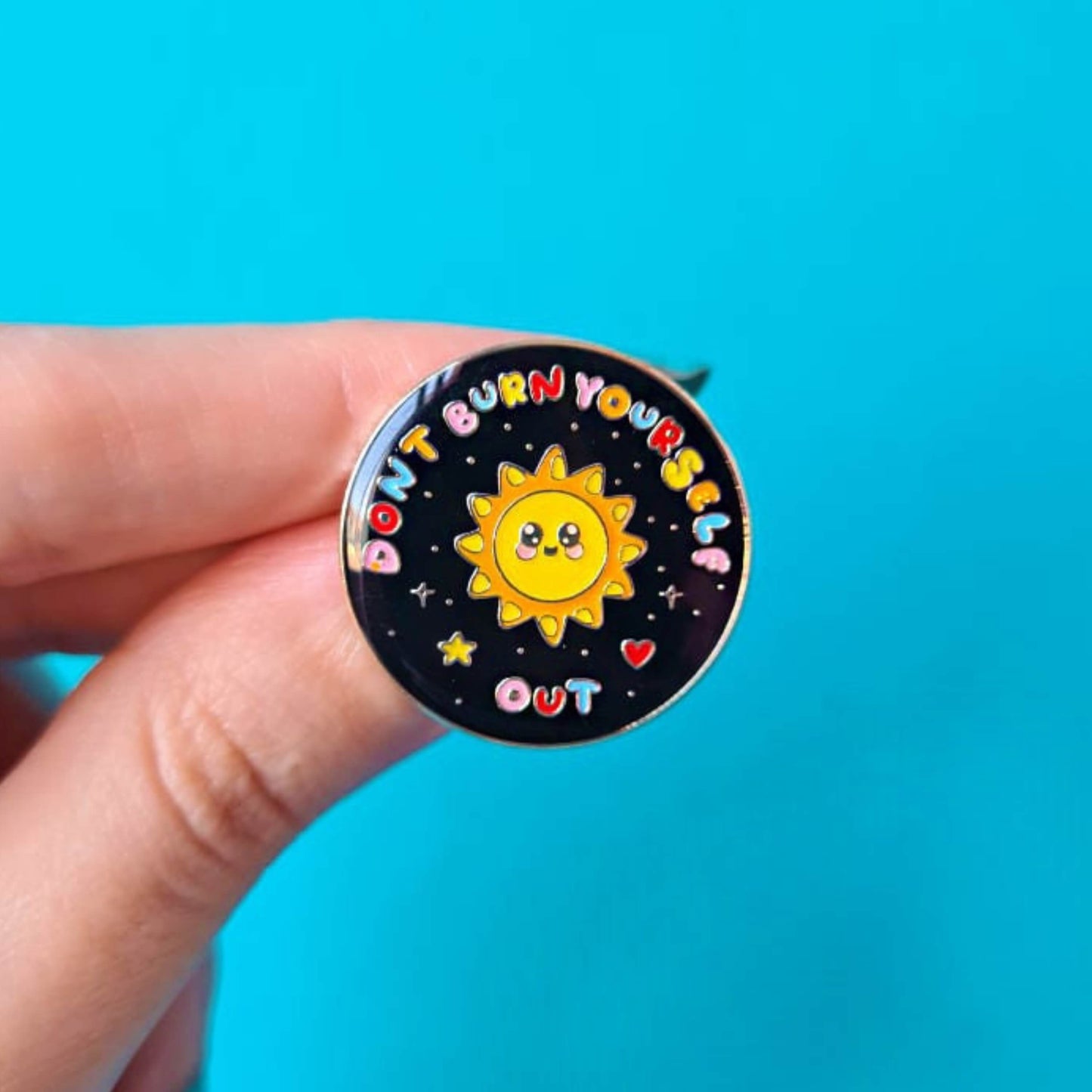 The Don't Burn Yourself Out Sunshine Enamel Pin being held over a blue background. The circle shaped pin badge is a black base with a yellow smiling sunshine in the centre with rainbow text surrounding it reading 'don't burn yourself out' along with a red heart, yellow star and silver sparkles. The design was created as a gentle reminder to practise self care.