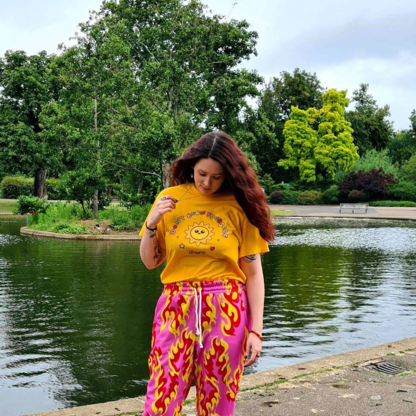 The Don't Burn Yourself Out Tee modelled by the innabox owner, Nikky, who has brown long hair and pink flame joggers. She is stood outside smiling looking down in front of a large pond with greenery. The yellow base cotton tshirt features a smiling happy sunshine with red, yellow and blue polka dots, a red heart, a yellow star and black sparkles surrounding it. Around the sunshine in rainbow bubble writing reads 'don't burn yourself out'. The design is a gentle reminder to practise self care.