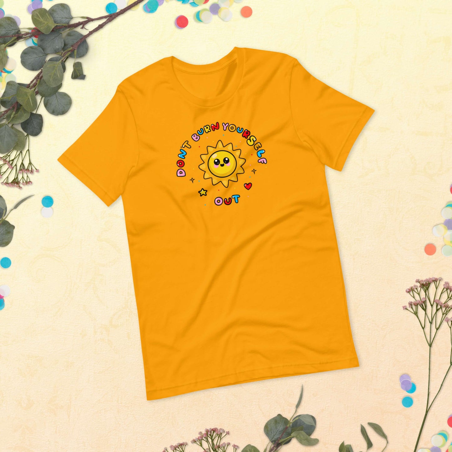 The Don't Burn Yourself Out Tee laying on a pastel yellow background with green foliage and multicoloured confetti circles. The yellow base cotton tshirt features a smiling happy sunshine with red, yellow and blue polka dots, a red heart, a yellow star and black sparkles surrounding it. Around the sunshine in rainbow bubble writing reads 'don't burn yourself out'. The design is a gentle reminder to practise self care.