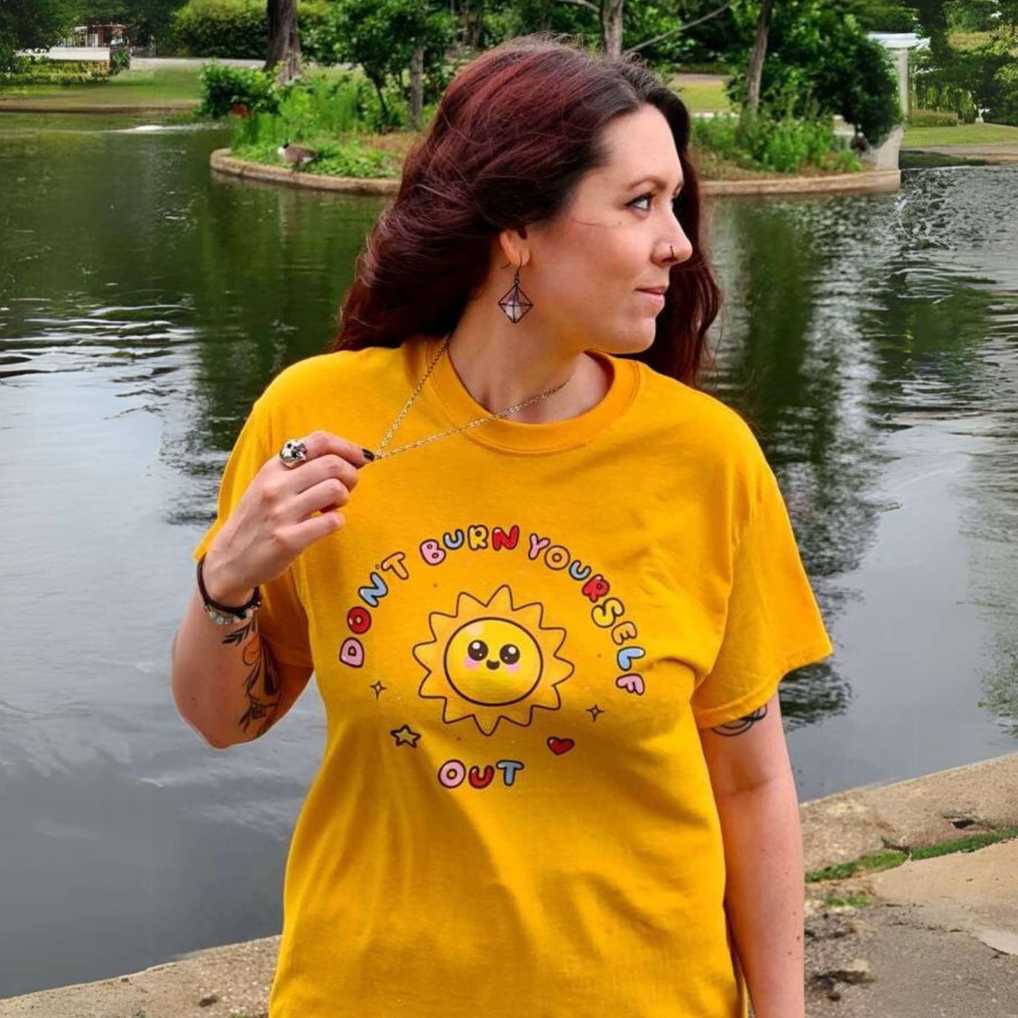The Don't Burn Yourself Out Tee modelled by the innabox owner, Nikky, who has brown long hair and tattoos. She is stood outside smiling looking off to the right in front of a large pond with greenery. The yellow base cotton tshirt features a smiling happy sunshine with red, yellow and blue polka dots, a red heart, a yellow star and black sparkles surrounding it. Around the sunshine in rainbow bubble writing reads 'don't burn yourself out'. The design is a gentle reminder to practise self care.
