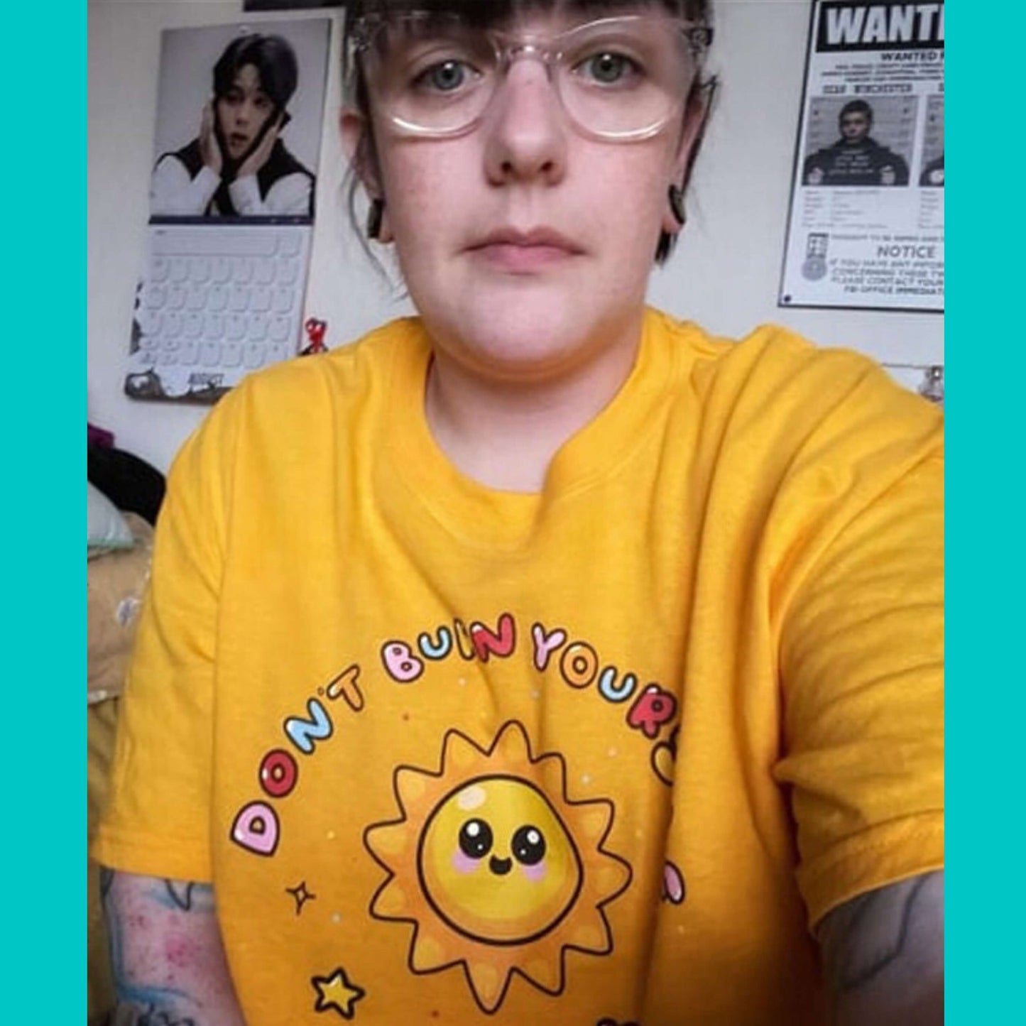 The Don't Burn Yourself Out Tee being worn cropped by a customer taking a selfie in their bedroom. The yellow base cotton tshirt features a smiling happy sunshine with red, yellow and blue polka dots, a red heart, a yellow star and black sparkles surrounding it. Around the sunshine in rainbow bubble writing reads 'don't burn yourself out'. The design is a gentle reminder to practise self care.
