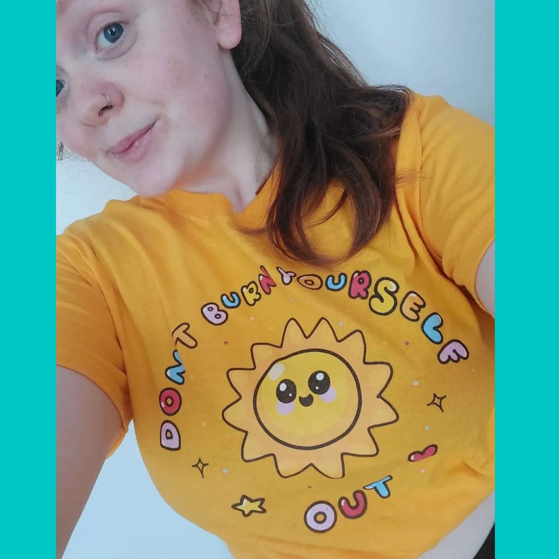 The Don't Burn Yourself Out Tee being worn cropped by a smiling customer taking a selfie in front of a white wall. The yellow base cotton tshirt features a smiling happy sunshine with red, yellow and blue polka dots, a red heart, a yellow star and black sparkles surrounding it. Around the sunshine in rainbow bubble writing reads 'don't burn yourself out'. The design is a gentle reminder to practise self care.