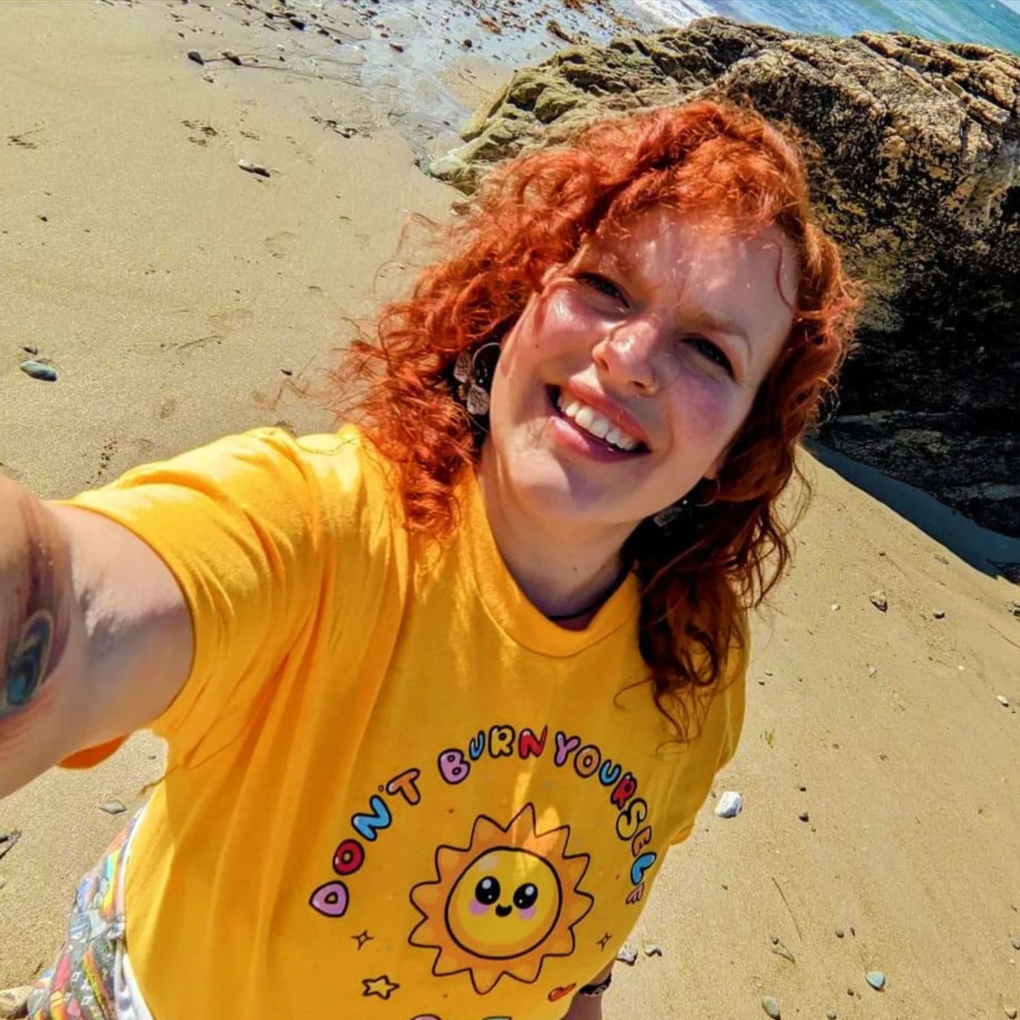 The Don't Burn Yourself Out Tee being worn by a smiling customer with red curly hair taking a selfie on a sandy sunny beach. The yellow base cotton tshirt features a smiling happy sunshine with red, yellow and blue polka dots, a red heart, a yellow star and black sparkles surrounding it. Around the sunshine in rainbow bubble writing reads 'don't burn yourself out'. The design is a gentle reminder to practise self care.