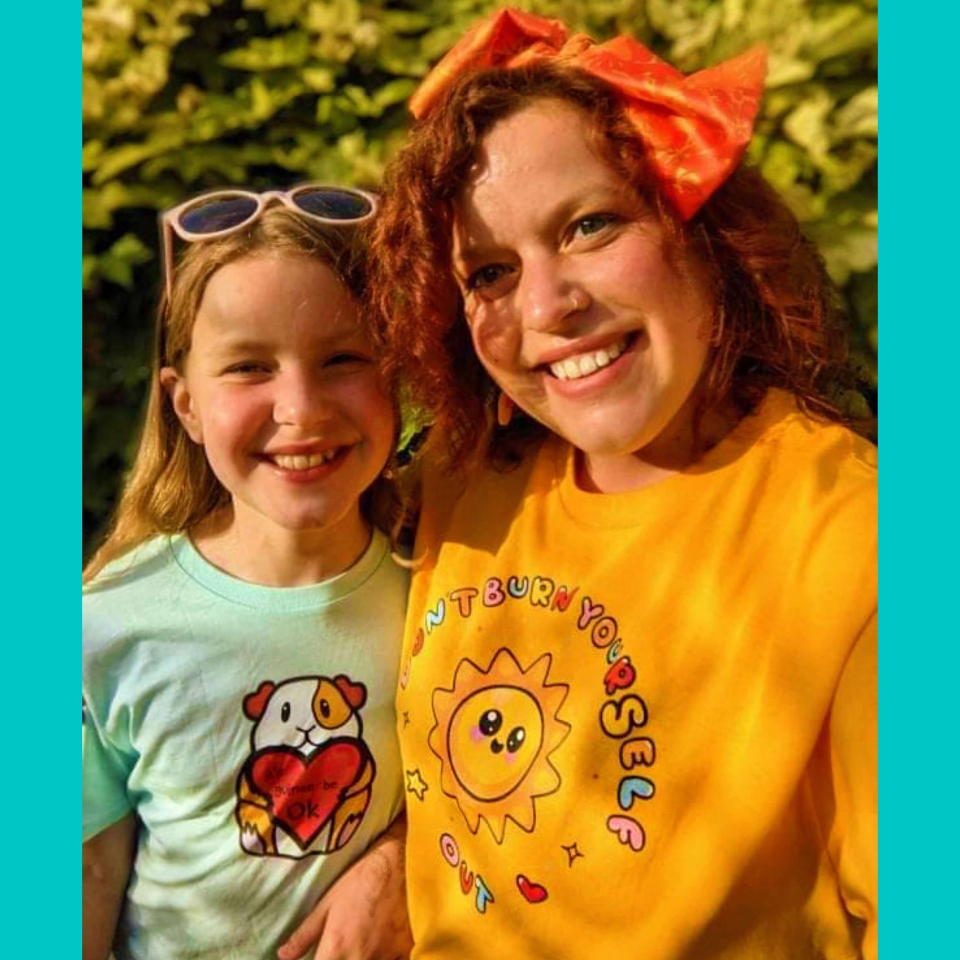 The Don't Burn Yourself Out Tee being worn by a smiling customer with red curly hair taking a selfie outside with their daughter wearing the guinea be ok tshirt from innabox. The yellow base cotton tshirt features a smiling happy sunshine with red, yellow and blue polka dots, a red heart, a yellow star and black sparkles surrounding it. Around the sunshine in rainbow bubble writing reads 'don't burn yourself out'. The design is a gentle reminder to practise self care.