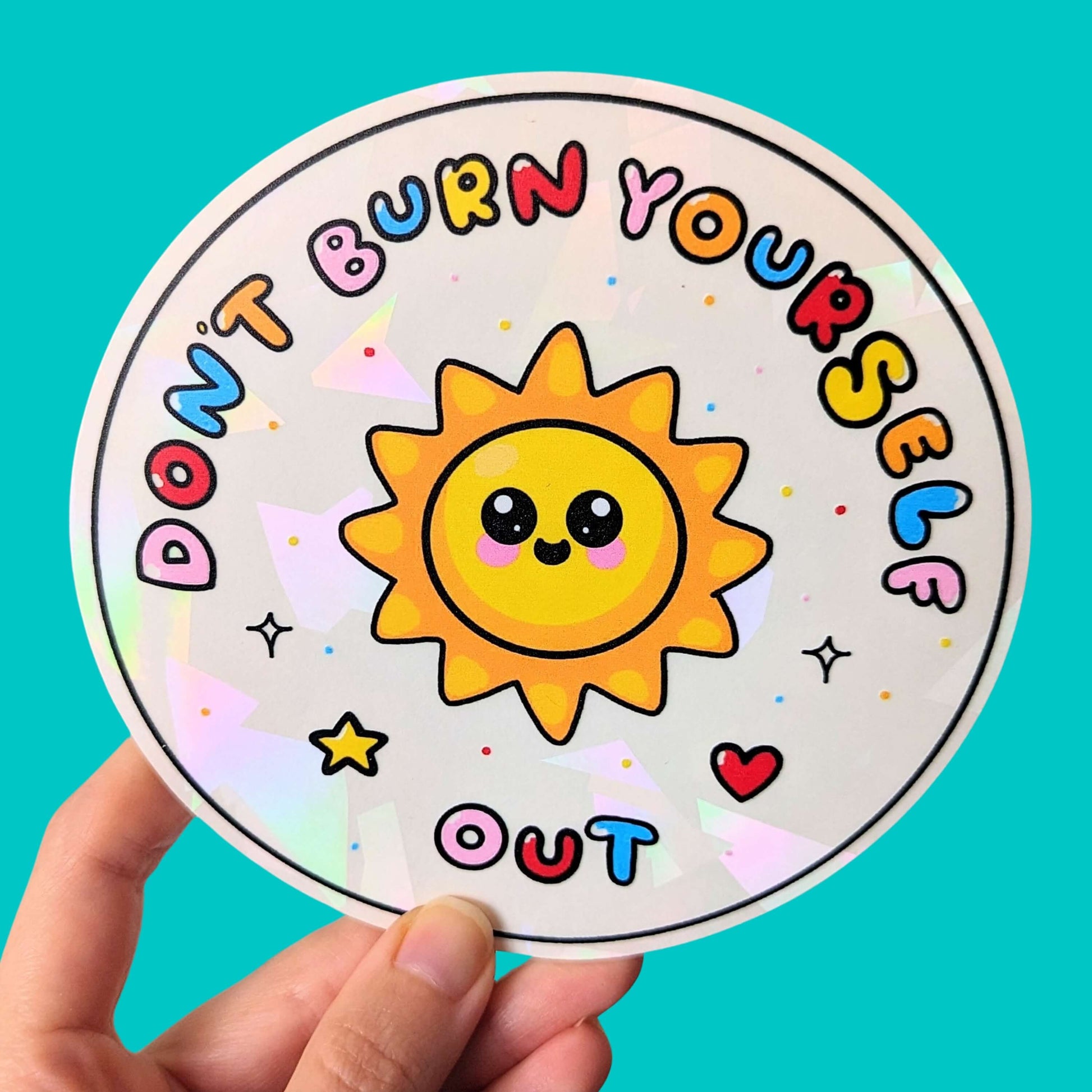 The Don't Burn Yourself Out Sun Catcher Rainbow Window Sticker being held up in front of a blue background. The circle holographic sticker features a smiling sunshine with pink blush in the middle with rainbow bubble writing reading 'don't burn yourself out' with a yellow star, red heart, black sparkle outlines and rainbow dots all over. The design is inspired by self care.