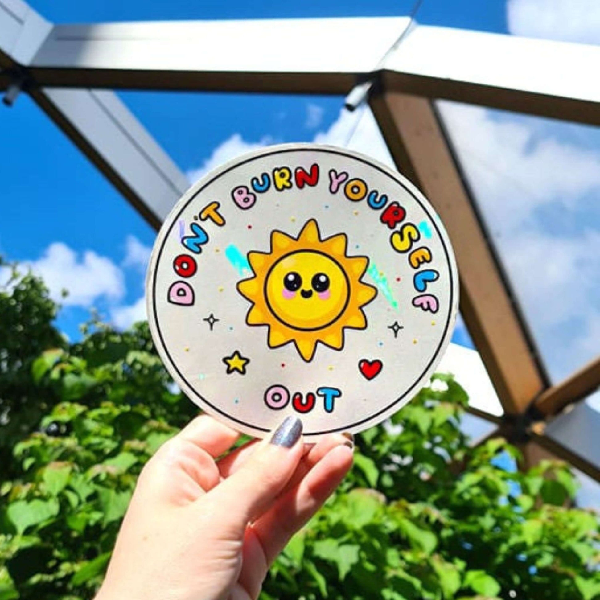 The Don't Burn Yourself Out Sun Catcher Rainbow Window Sticker being held up in front of an outdoor greenhouse full of green plants. The circle holographic sticker features a smiling sunshine with pink blush in the middle with rainbow bubble writing reading 'don't burn yourself out' with a yellow star, red heart, black sparkle outlines and rainbow dots all over. The design is inspired by self care.
