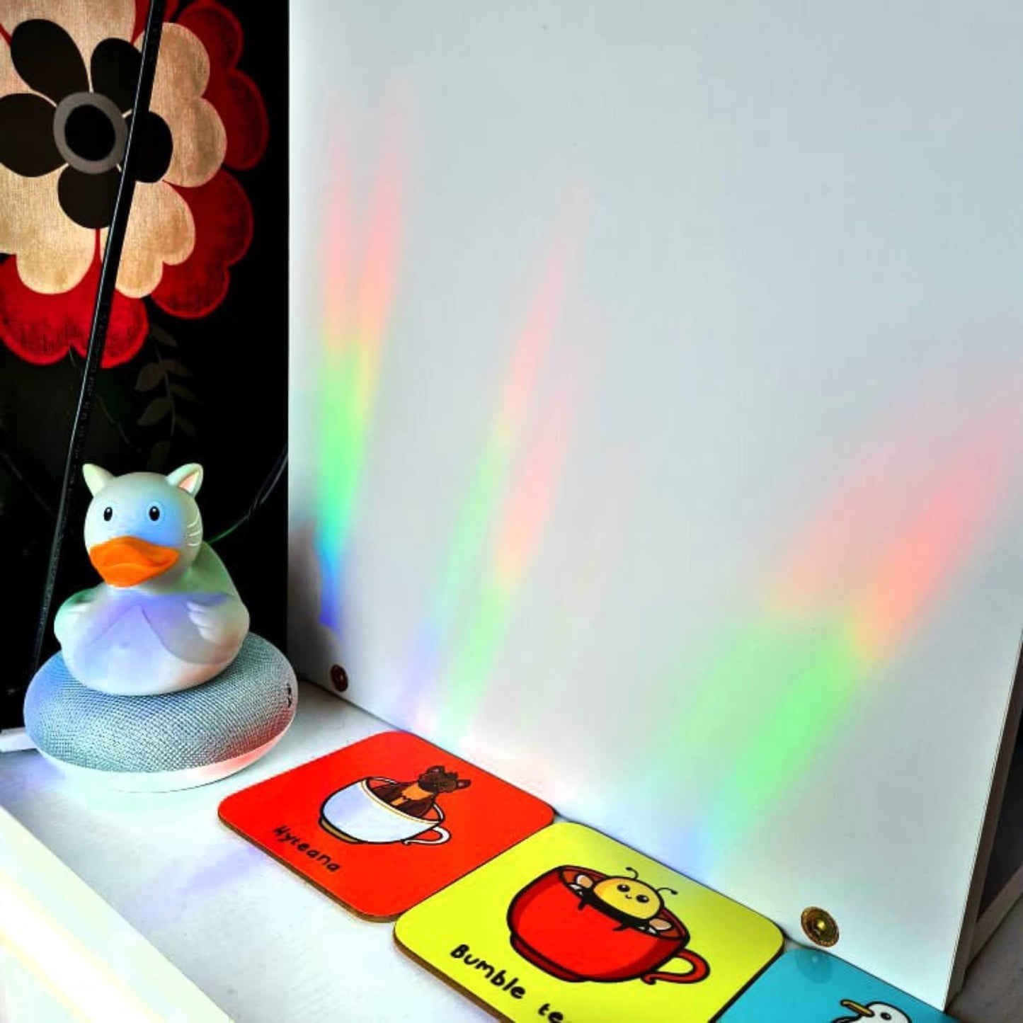 Rainbows being cast all over a white desk and storage unit of innabox coasters and a rubber cat duck from the Don't Burn Yourself Out Sun Catcher Rainbow Window Sticker. The holographic design is inspired by self care.