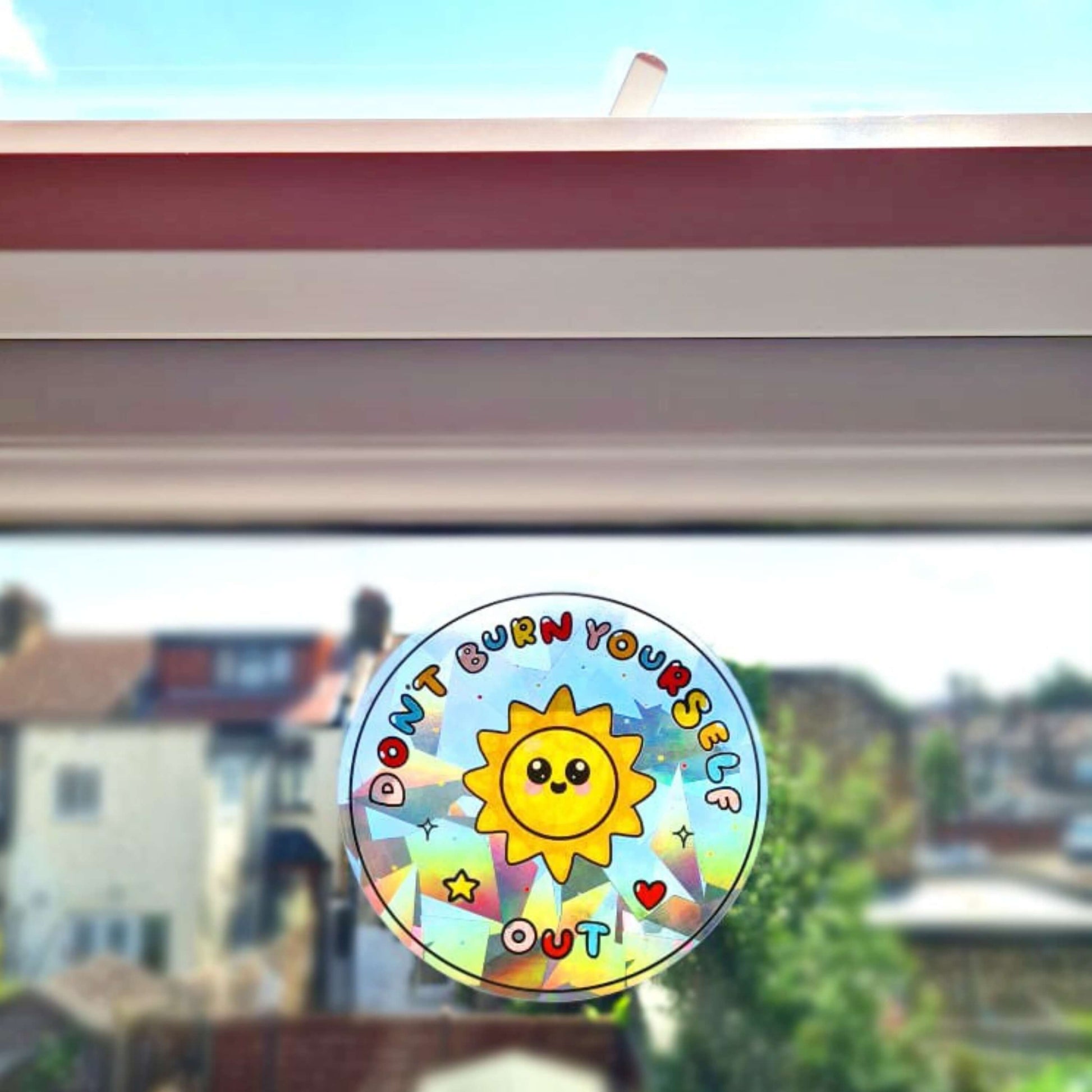 The Don't Burn Yourself Out Sun Catcher Rainbow Window Sticker stuck on a window with blue skies and houses behind. The circle holographic sticker features a smiling sunshine with pink blush in the middle with rainbow bubble writing reading 'don't burn yourself out' with a yellow star, red heart, black sparkle outlines and rainbow dots all over. The design is inspired by self care.