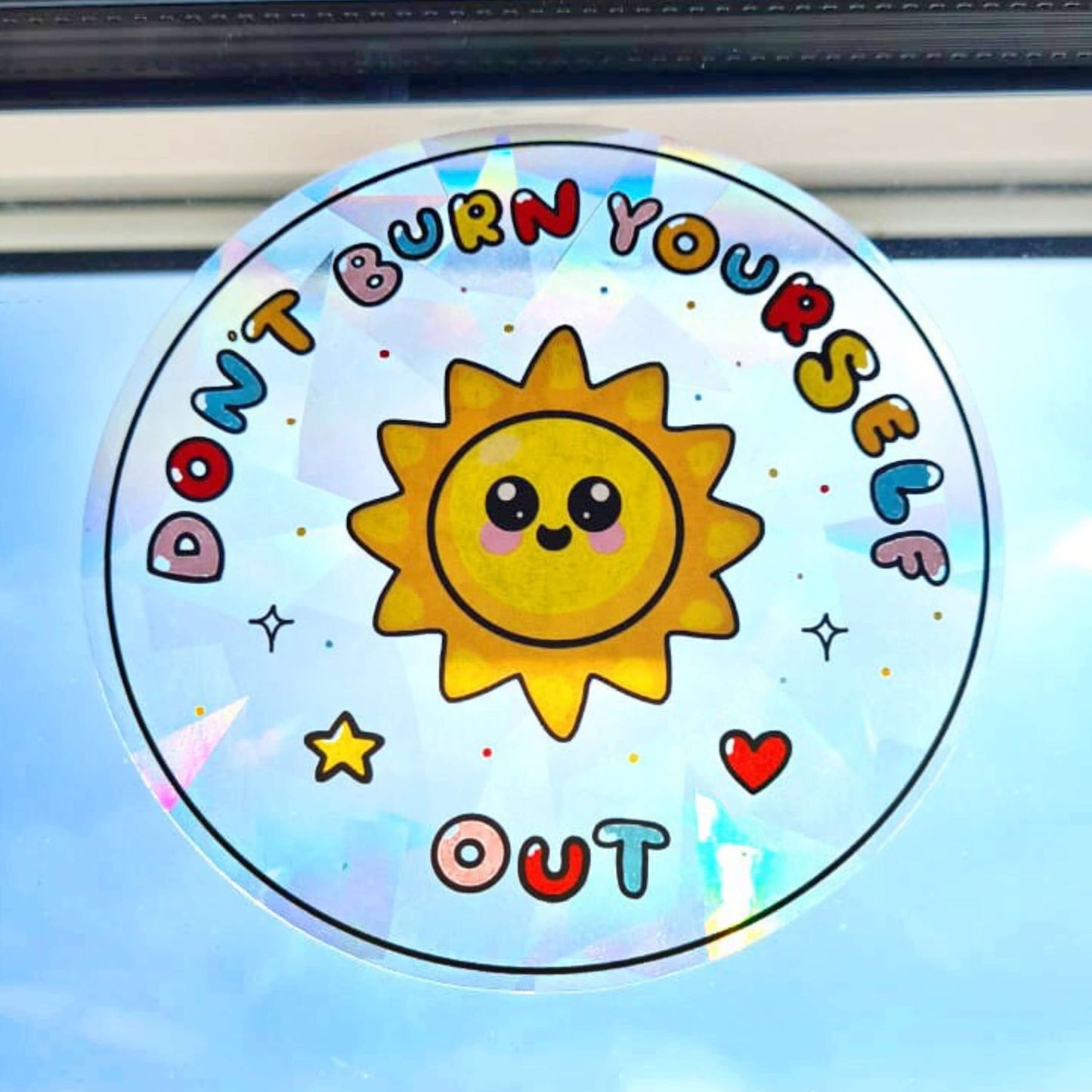 The Don't Burn Yourself Out Sun Catcher Rainbow Window Sticker stuck on a window with blue skies behind. The circle holographic sticker features a smiling sunshine with pink blush in the middle with rainbow bubble writing reading 'don't burn yourself out' with a yellow star, red heart, black sparkle outlines and rainbow dots all over. The design is inspired by self care.