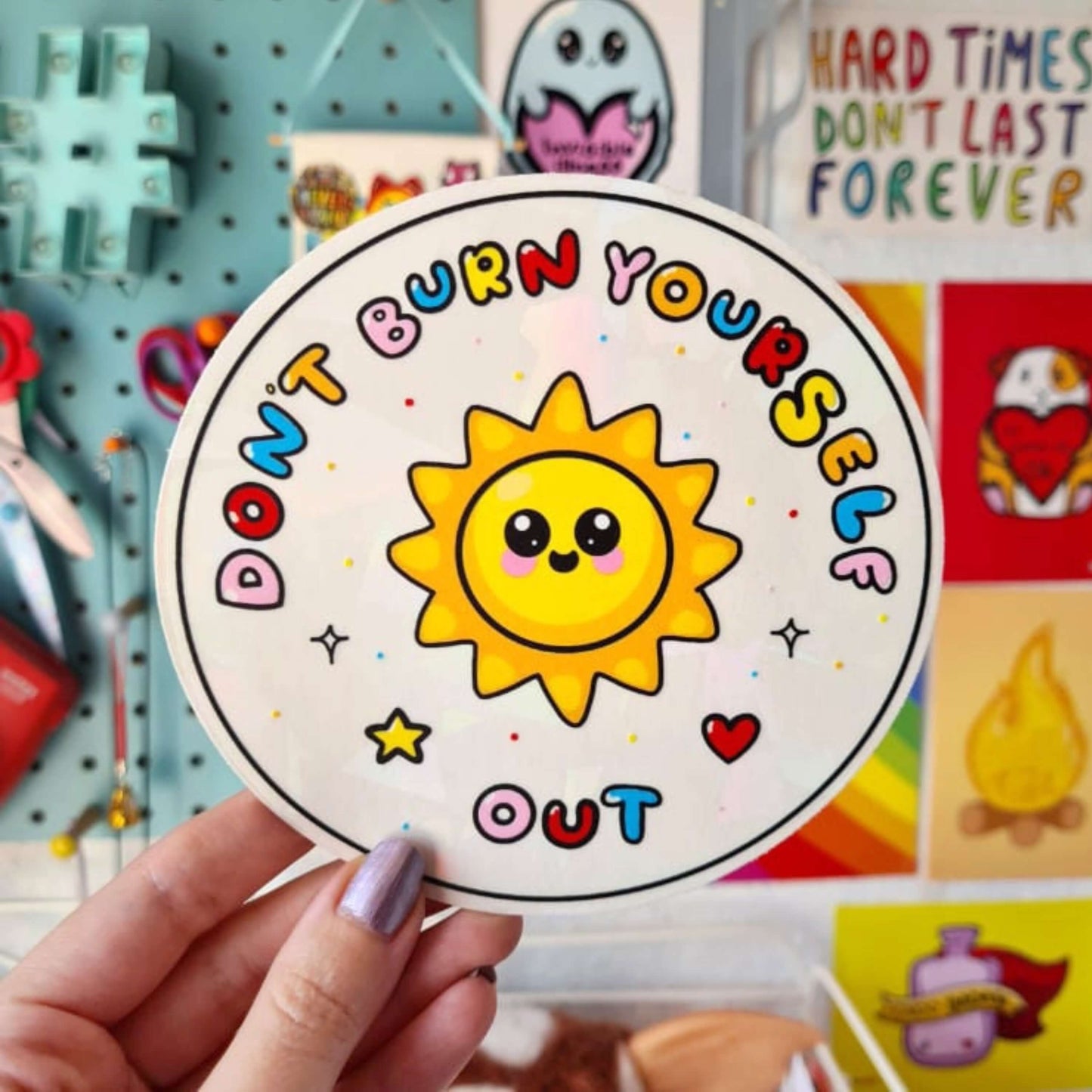 The Don't Burn Yourself Out Sun Catcher Rainbow Window Sticker being held up in front of a blue pinboard with other innabox products. The circle holographic sticker features a smiling sunshine with pink blush in the middle with rainbow bubble writing reading 'don't burn yourself out' with a yellow star, red heart, black sparkle outlines and rainbow dots all over. The design is inspired by self care.