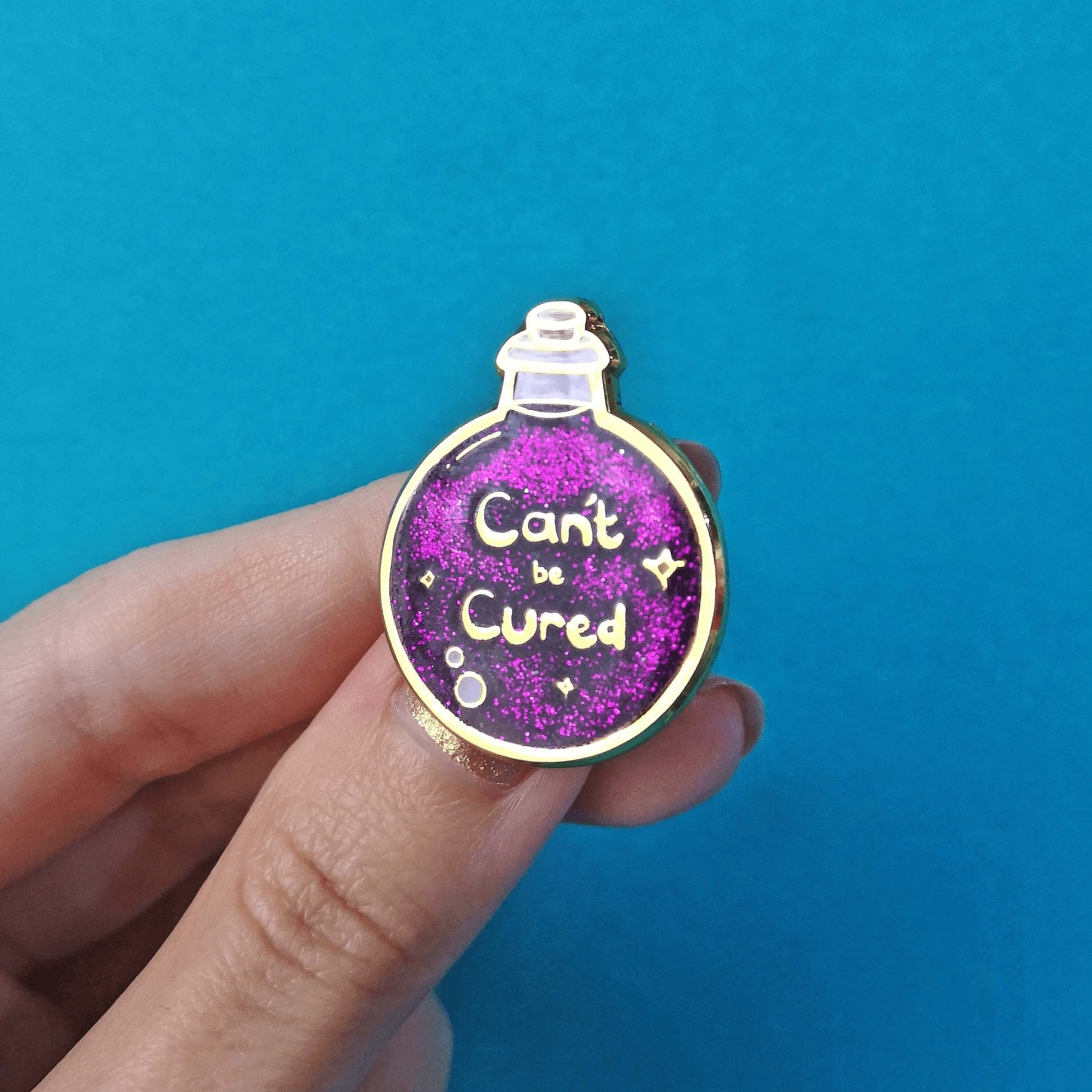 The Can't Be Cured Potion Bottle Enamel Pin being held over a blue background by a hand with gold glitter nail varnish. A gold outlined circular potion bottle with a glittery purple middle with two pastel pink bubbles, gold sparkles and gold text that reads 'can't be cured'. The design was inspired by chronic illnesses.