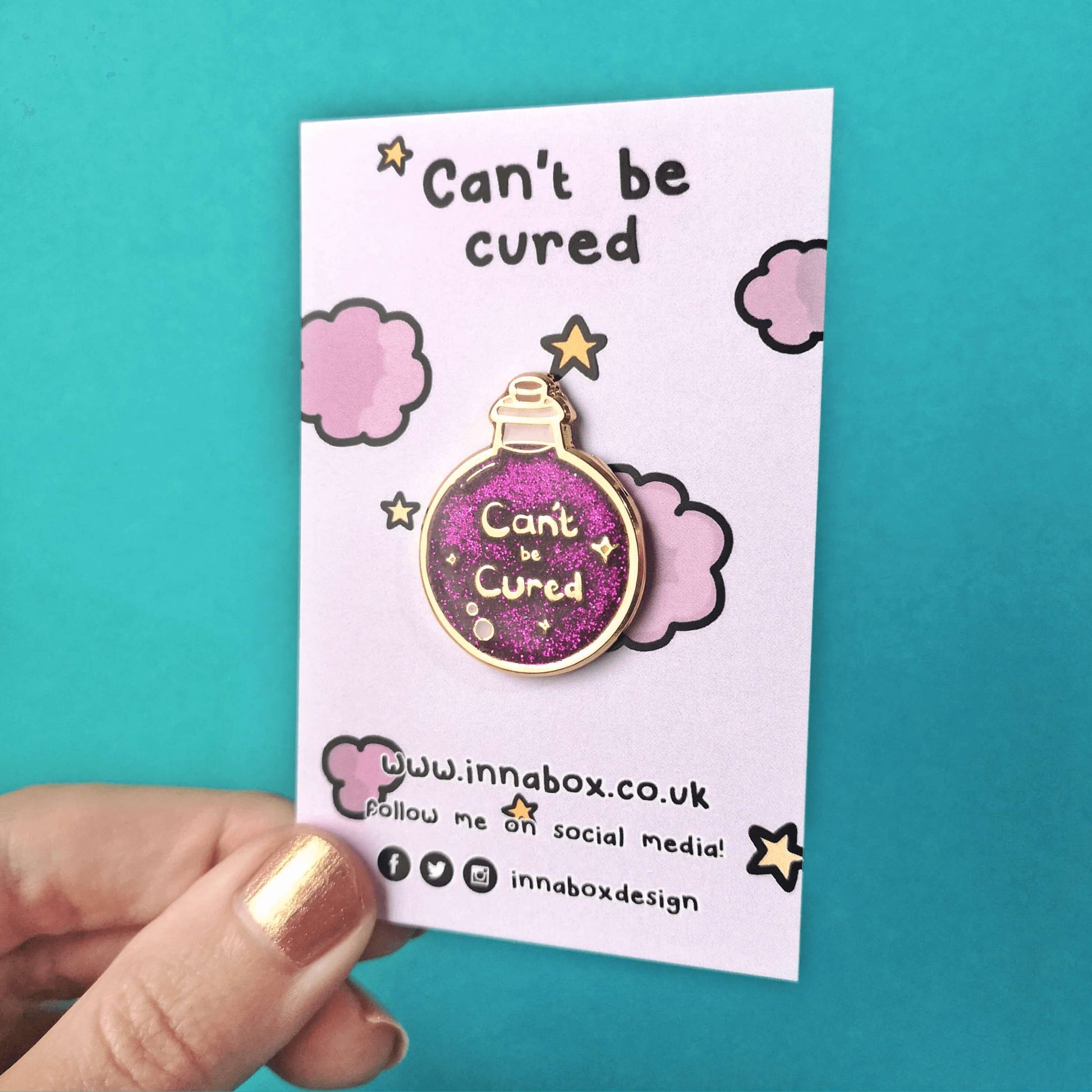 The Can't Be Cured Potion Bottle Enamel Pin on a lilac backing card with purple clouds and yellow stars, with can't be cured written at the top and innabox social media handles written at the bottom both in black, being held over a blue background. The pin is a gold outlined circular potion bottle with a glittery purple middle with two pastel pink bubbles, gold sparkles and gold text that reads 'can't be cured'. The design was inspired by chronic illnesses.
