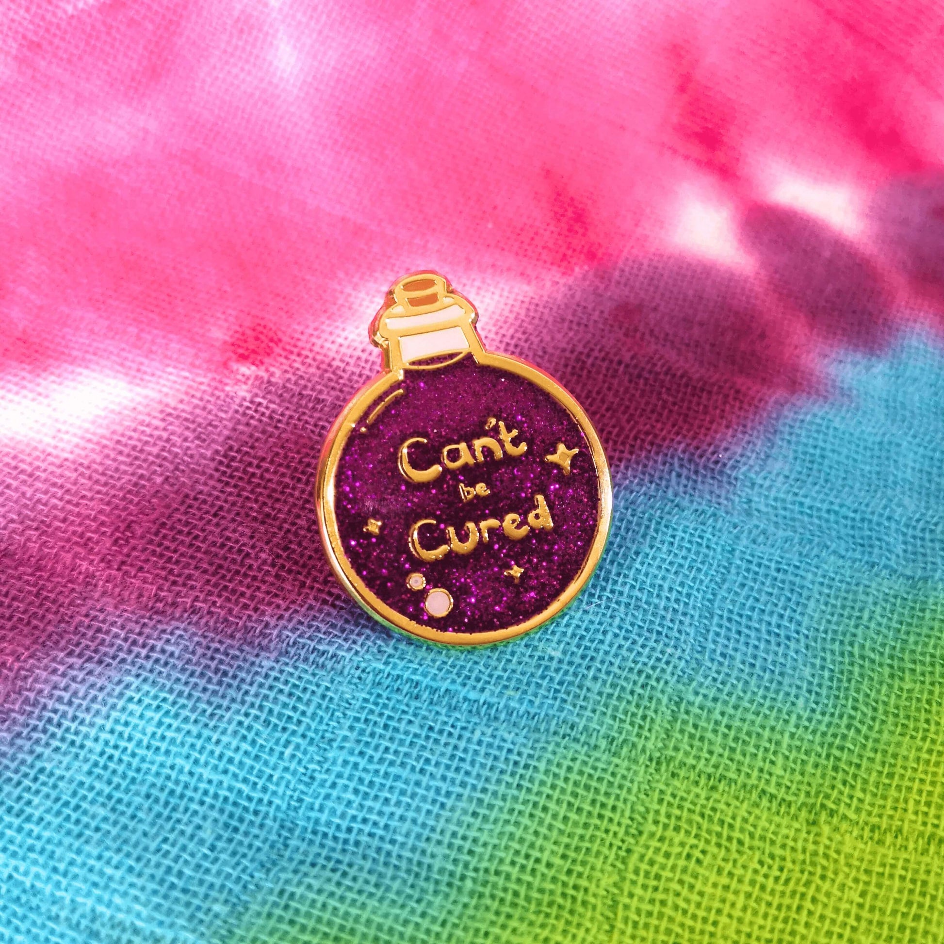 The Can't Be Cured Potion Bottle Enamel Pin on a rainbow tie dye fabric background. A gold outlined circular potion bottle with a glittery purple middle with two pastel pink bubbles, gold sparkles and gold text that reads 'can't be cured'. The design was inspired by chronic illnesses.