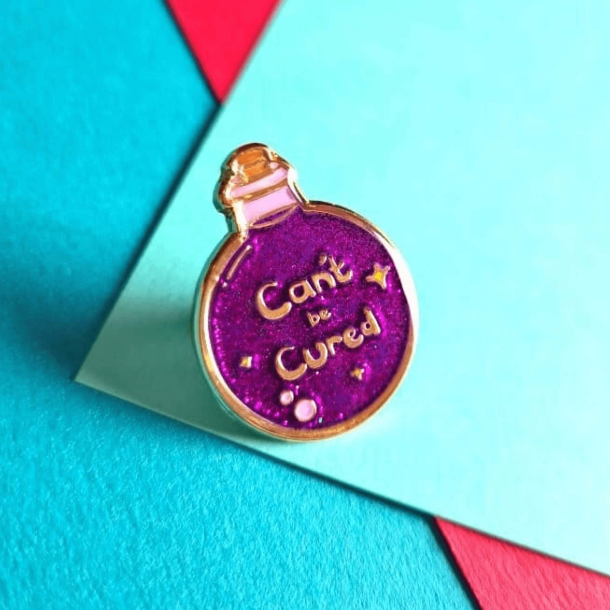 The Can't Be Cured Potion Bottle Enamel Pin on a blue, green and red background. A gold outlined circular potion bottle with a glittery purple middle with two pastel pink bubbles, gold sparkles and gold text that reads 'can't be cured'. The design was inspired by chronic illnesses.
