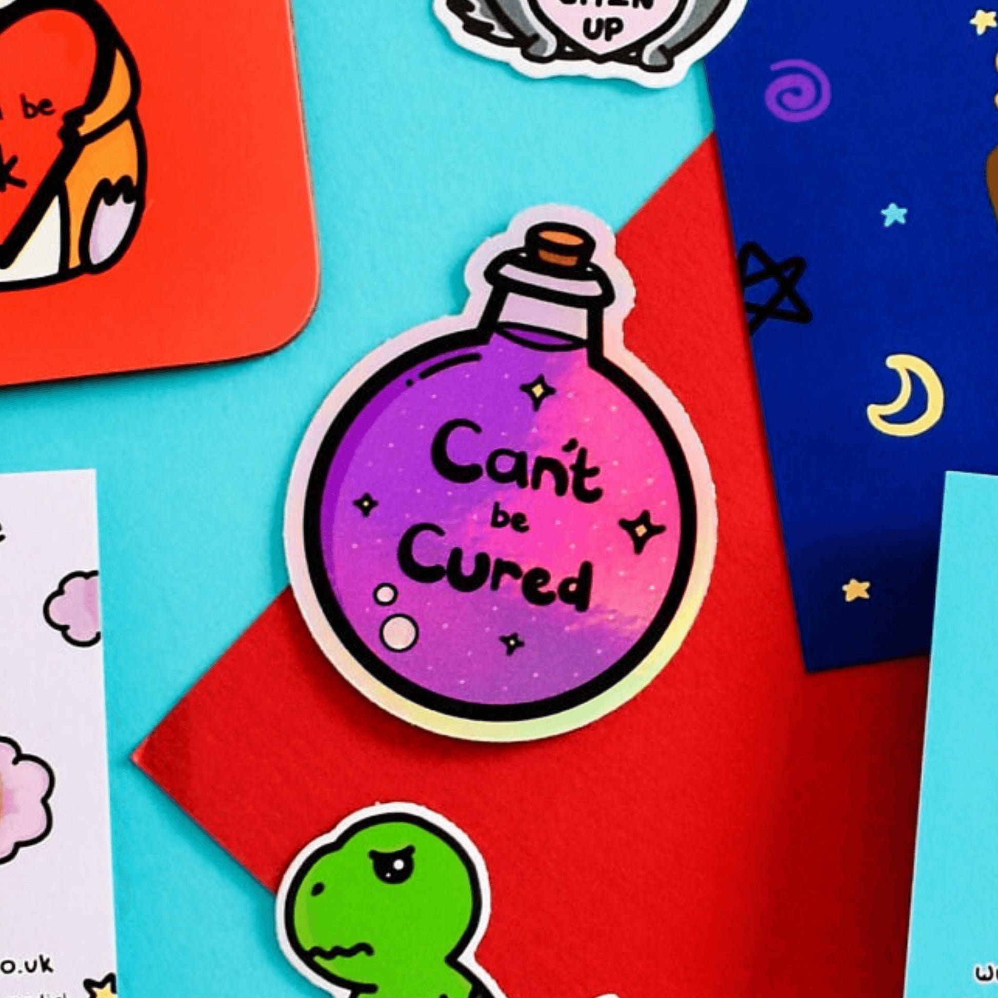 The Can't Be Cured Holographic Sticker on a red and blue background with various other innabox products surrounding it. The sticker is a circular potion bottle with a cork stop top and purple middle. In the middle is white and yellow sparkles, two pastel pink bubbles and black text that reads 'can't be cured'. The design was inspired by chronic illnesses.