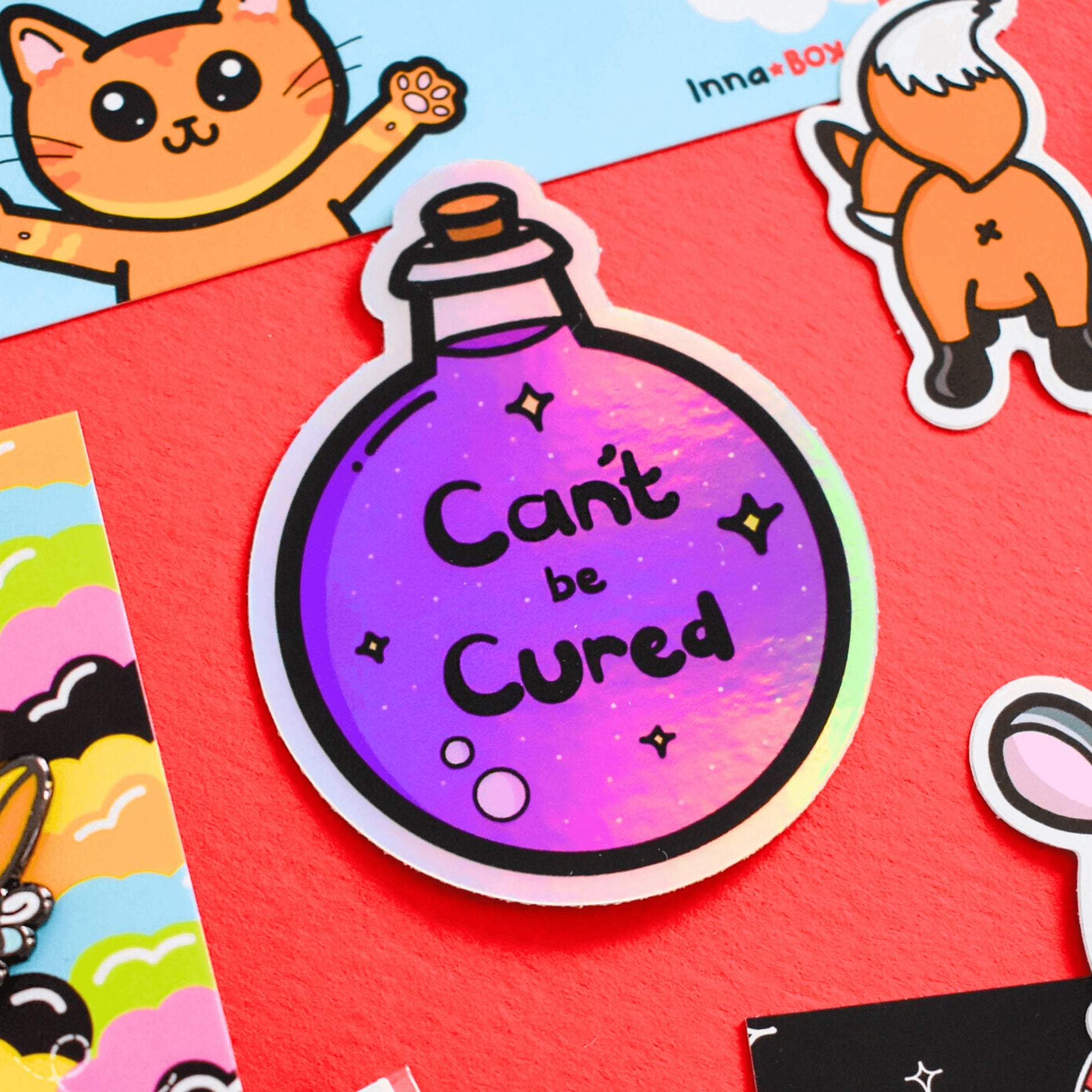 The Can't Be Cured Holographic Sticker on a red background with various other innabox products surrounding it. The sticker is a circular potion bottle with a cork stop top and purple middle. In the middle is white and yellow sparkles, two pastel pink bubbles and black text that reads 'can't be cured'. The design was inspired by chronic illnesses.