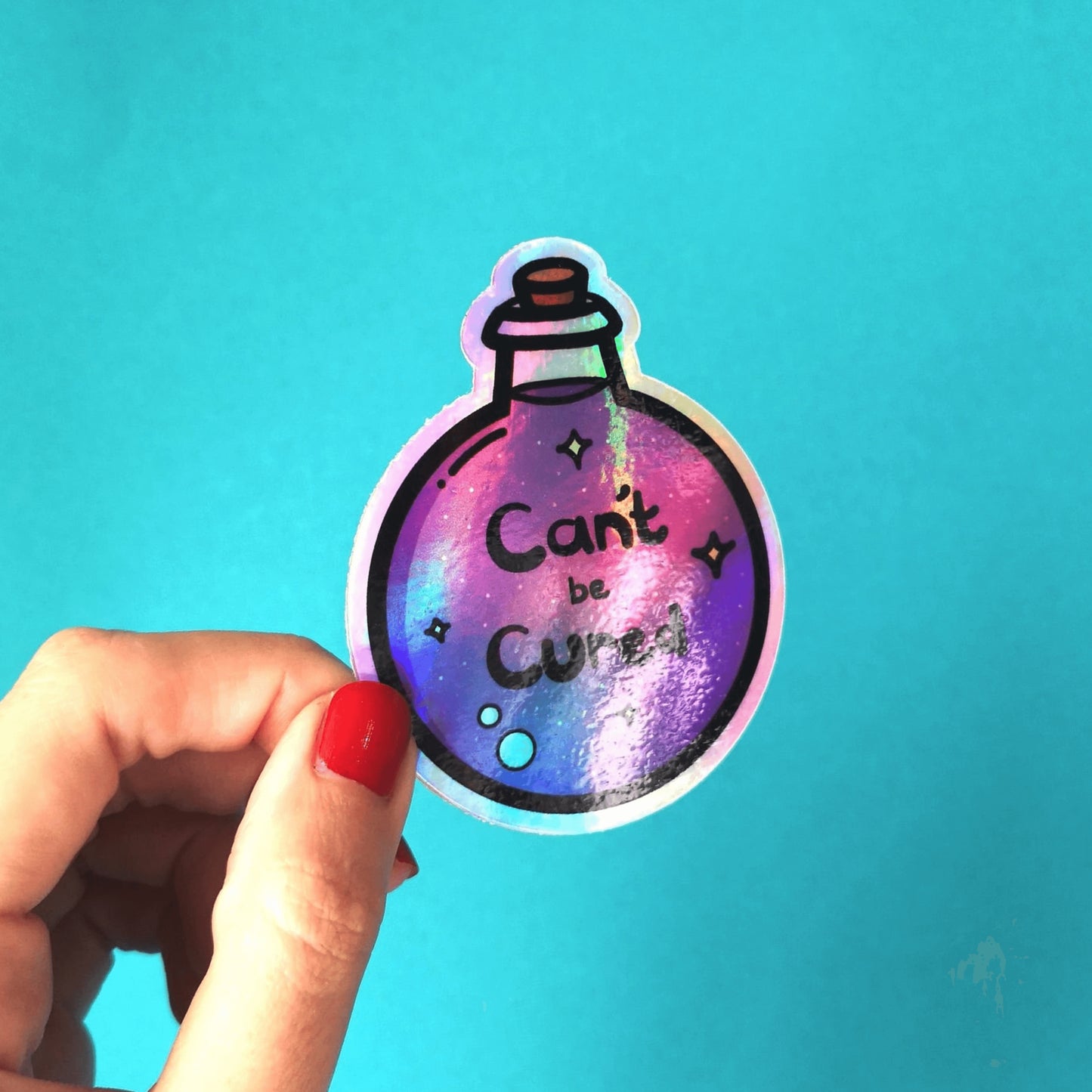 The Can't Be Cured Holographic Sticker being held over a blue background by a hand with red nail varnish. The sticker is a circular potion bottle with a cork stop top and purple middle. In the middle is white and yellow sparkles, two pastel pink bubbles and black text that reads 'can't be cured'. The design was inspired by chronic illnesses.