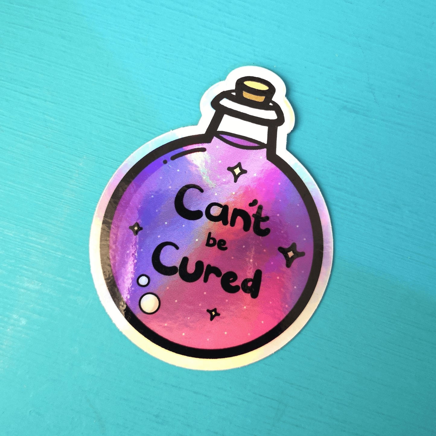 The Can't Be Cured Holographic Sticker on a blue background. The sticker is a circular potion bottle with a cork stop top and purple middle. In the middle is white and yellow sparkles, two pastel pink bubbles and black text that reads 'can't be cured'. The design was inspired by chronic illnesses.