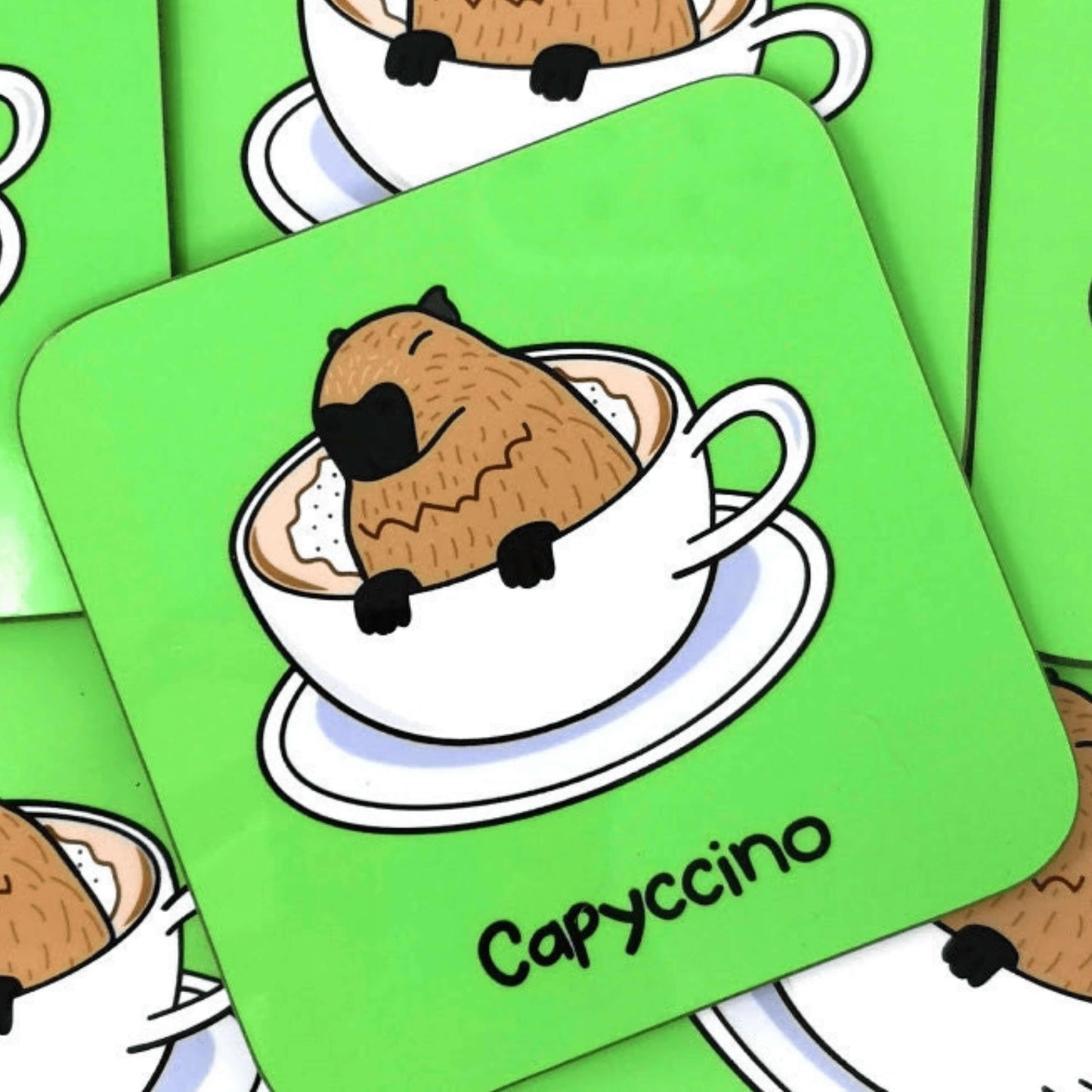 The Capyccino - Capybara Cappuccino Coaster on multiple other copies of the coaster. The wooden green coaster features a cute brown capybara sat peeking over a cappuccino in a white mug and saucer, underneath reads capyccino in black.