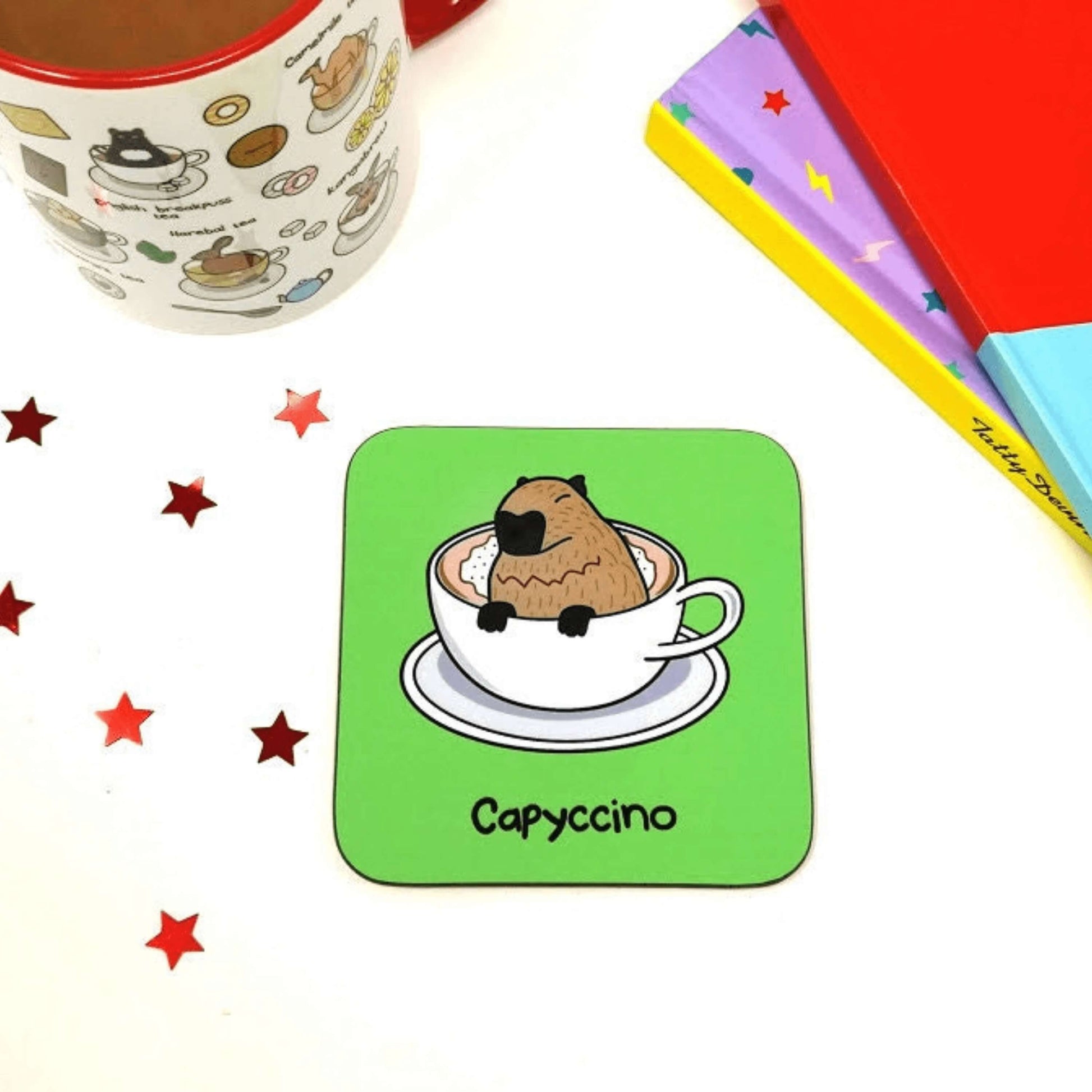 The Capyccino - Capybara Cappuccino Coaster on a white table with an innabox mug, red star sequins and colourful books. The wooden green coaster features a cute brown capybara sat peeking over a cappuccino in a white mug and saucer, underneath reads capyccino in black.