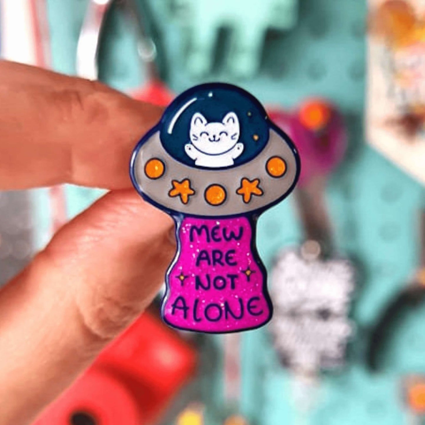 The Cat UFO Mew are not Alone Enamel Pin being held up with a blurry out of focus background. The blue outline pin is of a white smiling cat in a silver ufo spaceship with yellow glittery stars and circles, underneath is pink glittery beam with blue text reading 'mew are not alone' a pun on you are not alone.