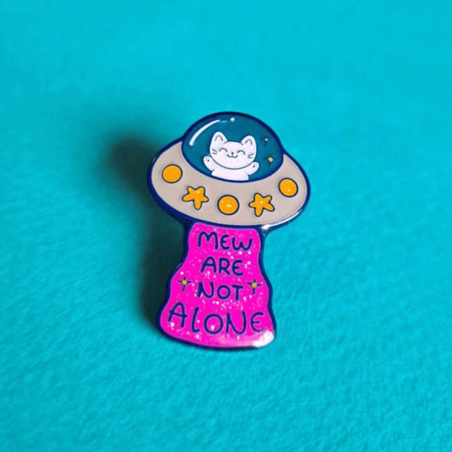 The Cat UFO Mew are not Alone Enamel Pin on a blue background. The blue outline pin is of a white smiling cat in a silver ufo spaceship with yellow glittery stars and circles, underneath is pink glittery beam with blue text reading 'mew are not alone' a pun on you are not alone.