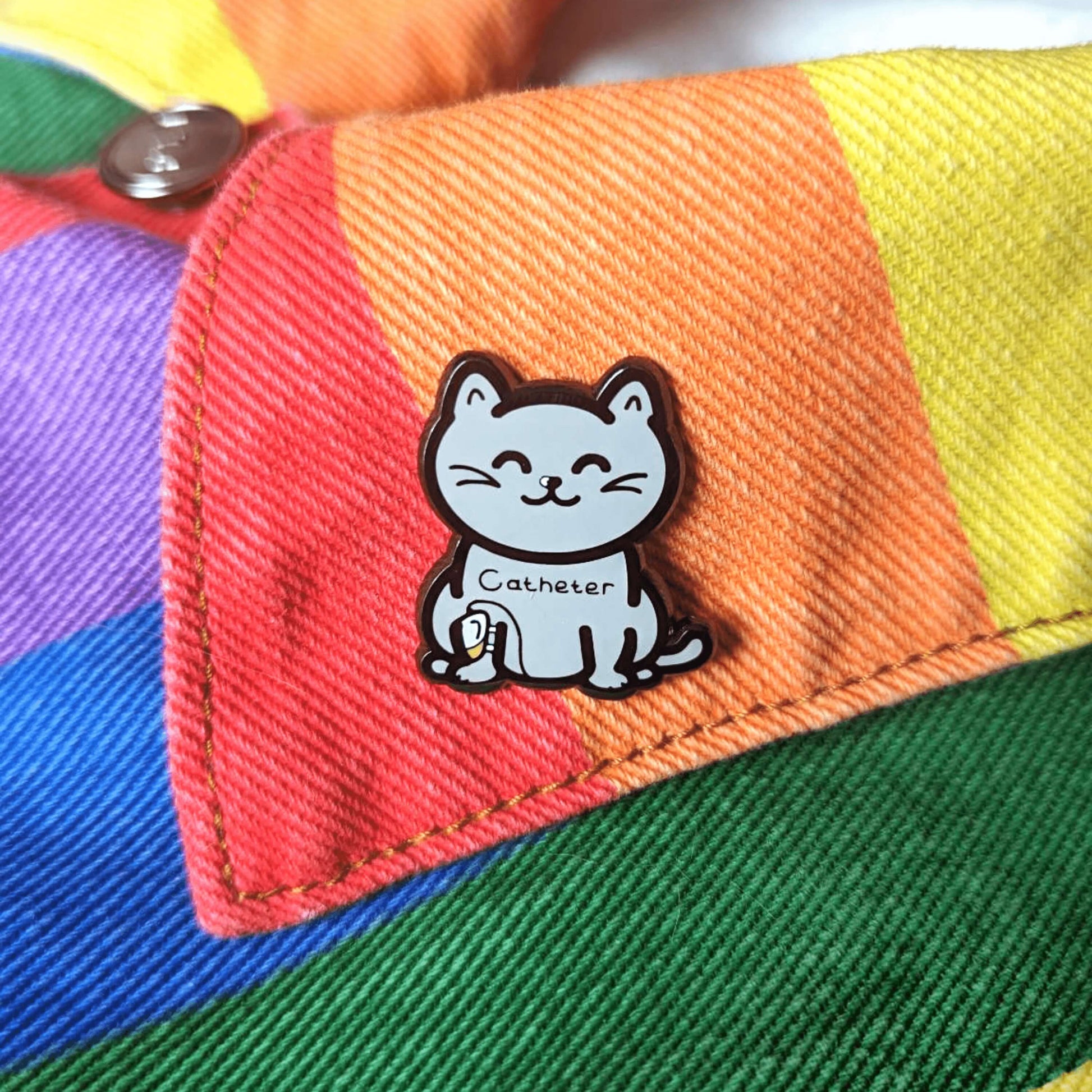 The Catheter Enamel Pin - Catheter on a rainbow denim jacket collar. The pin is a grey smiling cat sat down with a urine drainage Urostomy pouch strapped to its right leg and text across its chest reading 'catheter'. The pin is designed to raise awareness for bladder problems such as UTIs and other chronic illnesses.