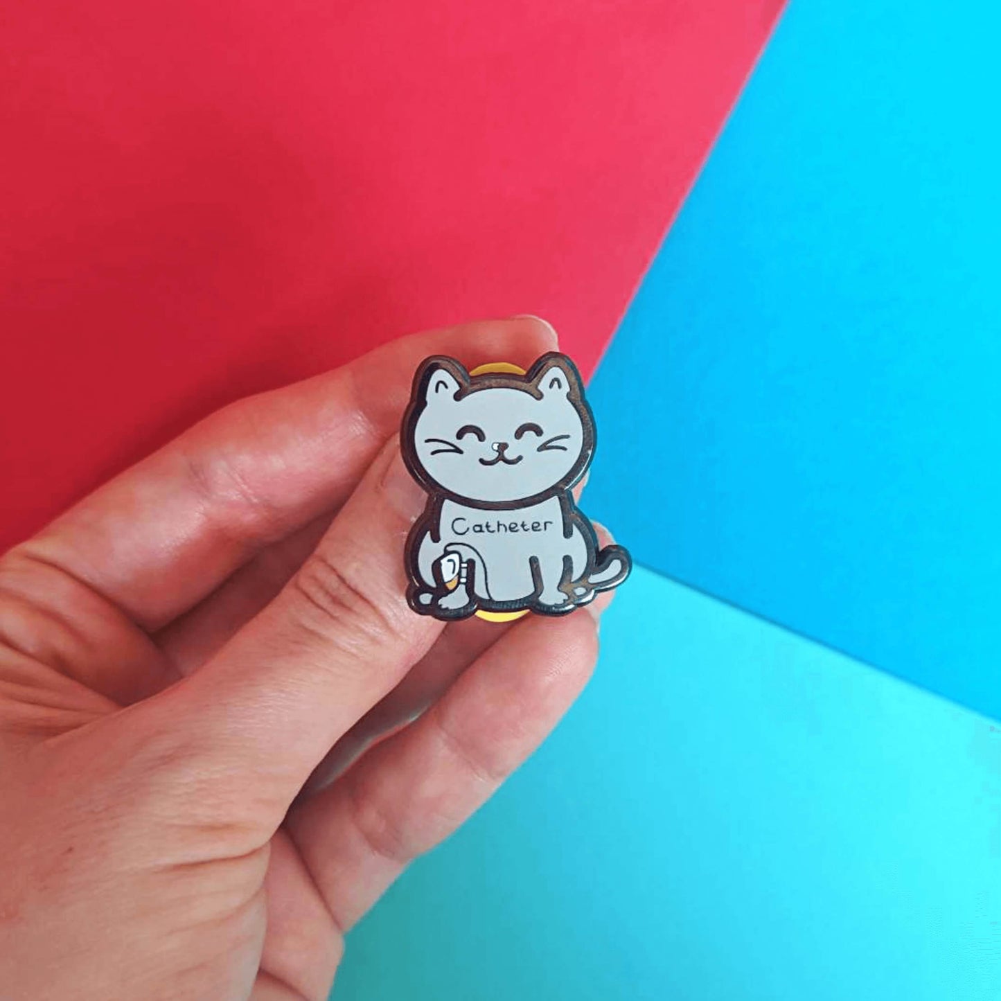 The Catheter Enamel Pin - Catheter being held over a red and blue background. The pin is a grey smiling cat sat down with a urine drainage Urostomy pouch strapped to its right leg and text across its chest reading 'catheter'. The pin is designed to raise awareness for bladder problems such as UTIs and other chronic illnesses.