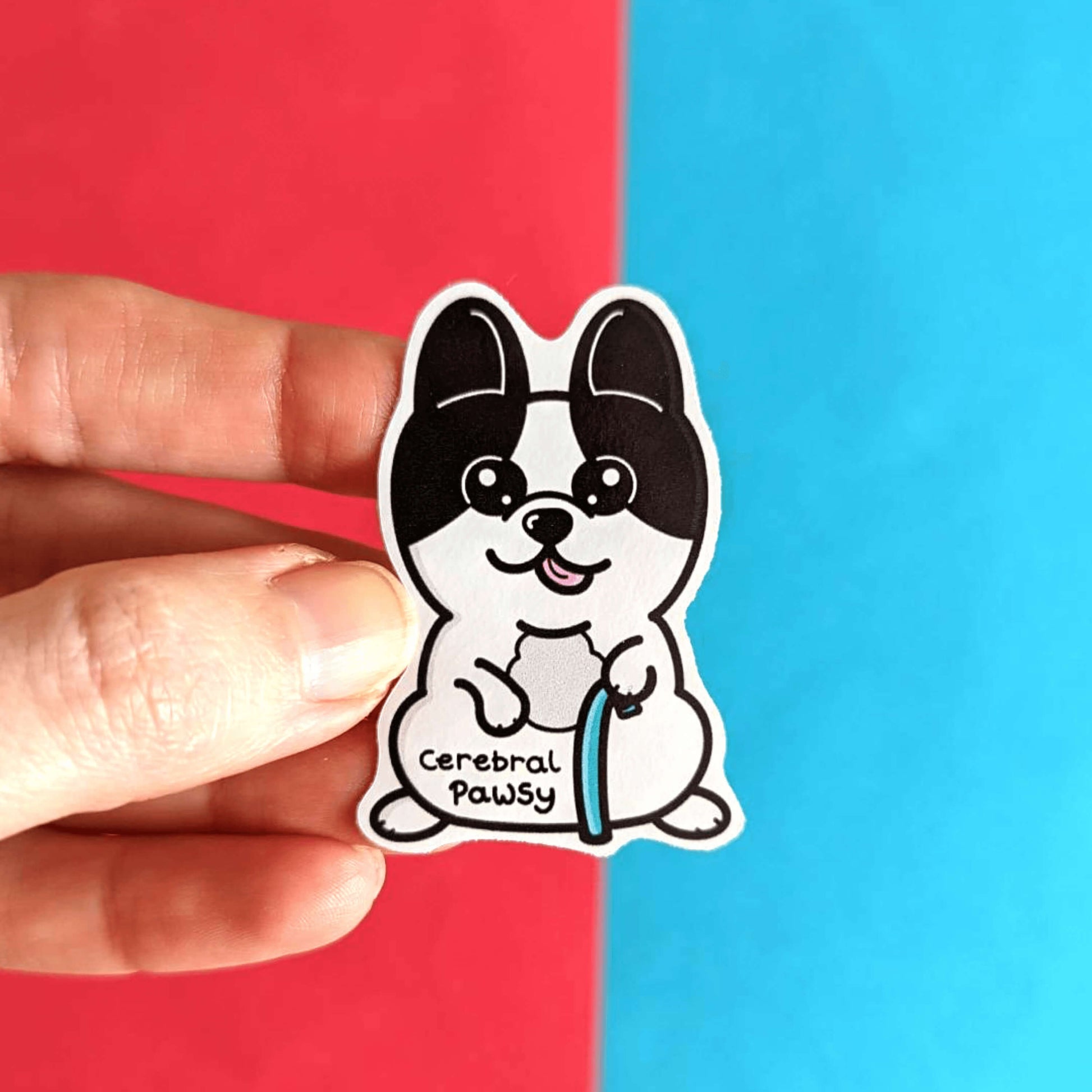 A hand holding the Cerebral Pawsy Sticker - Cerebral Palsy in front of a red and blue background. The sticker is a white and black dog with big eyes and pointy ears. The dog has it's pink tongue out and is standing on his back legs while holding a blue walking aid. 'Cerebral Pawsy' is written in black under it's tummy.