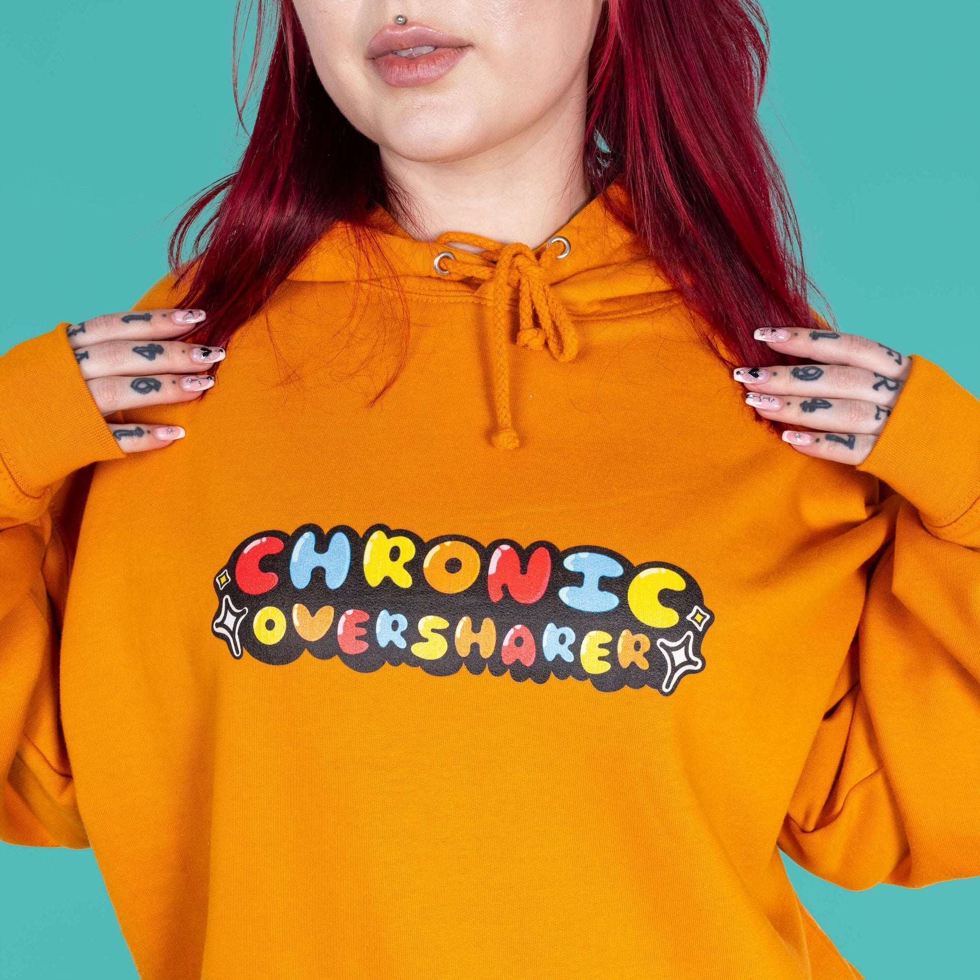 The Chronic Over Sharer Pumpkin Orange Hoodie modelled on Flo, a red haired alternative model, in front of a blue background. She is facing forward with both hands resting on her shoulders, the photo is cropped in on the middle. The pumpkin orange hoodie features a drawstring hood, a large front pocket and the text 'chronic oversharer' in rainbow bubble font with a black shadow effect and white sparkles. The design was created to raise awareness for neurodivergent disorders such as ADHD.