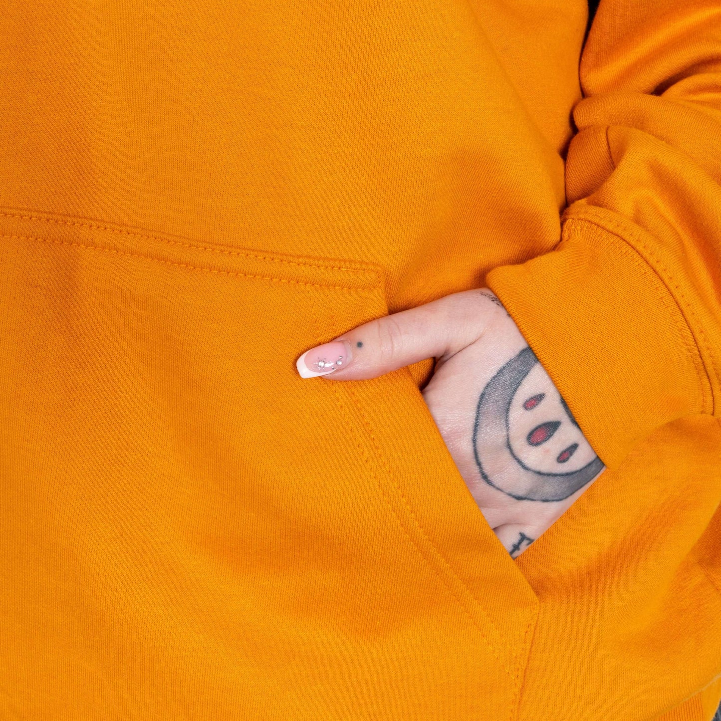 The Chronic Over Sharer Pumpkin Orange Hoodie modelled on Flo, a red haired alternative model, in front of a blue background. A close up of her tattooed hand in the front pocket. The pumpkin orange hoodie features a drawstring hood, a large front pocket and the text 'chronic oversharer' in rainbow bubble font with a black shadow effect and white sparkles. The design was created to raise awareness for neurodivergent disorders such as ADHD.