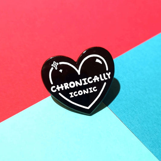 The Chronically Iconic Enamel Pin on a red and blue background. The black heart shaped pin has a white outline with sparkles and text reading 'chronically iconic'. The design was created to raise awareness for chronic illness and invisible illness.