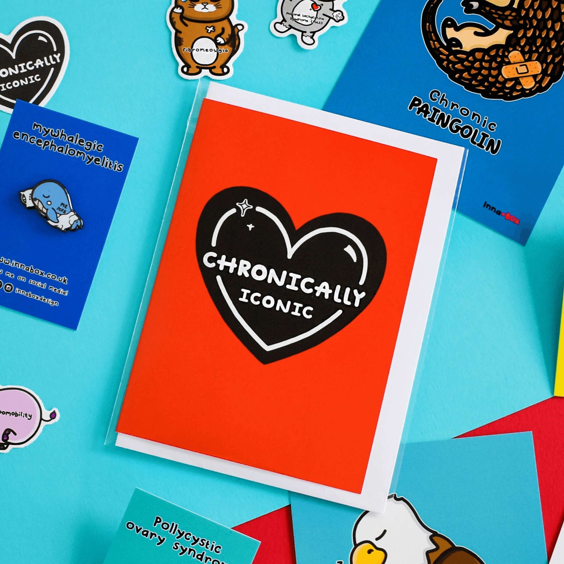 The Chronically Iconic Card on a blue background with other innabox products and a white envelope underneath. The red a6 card features a black heart with a white outline, sparkles and text that reads 'chronically iconic'. The design is to raise awareness for chronic illness.