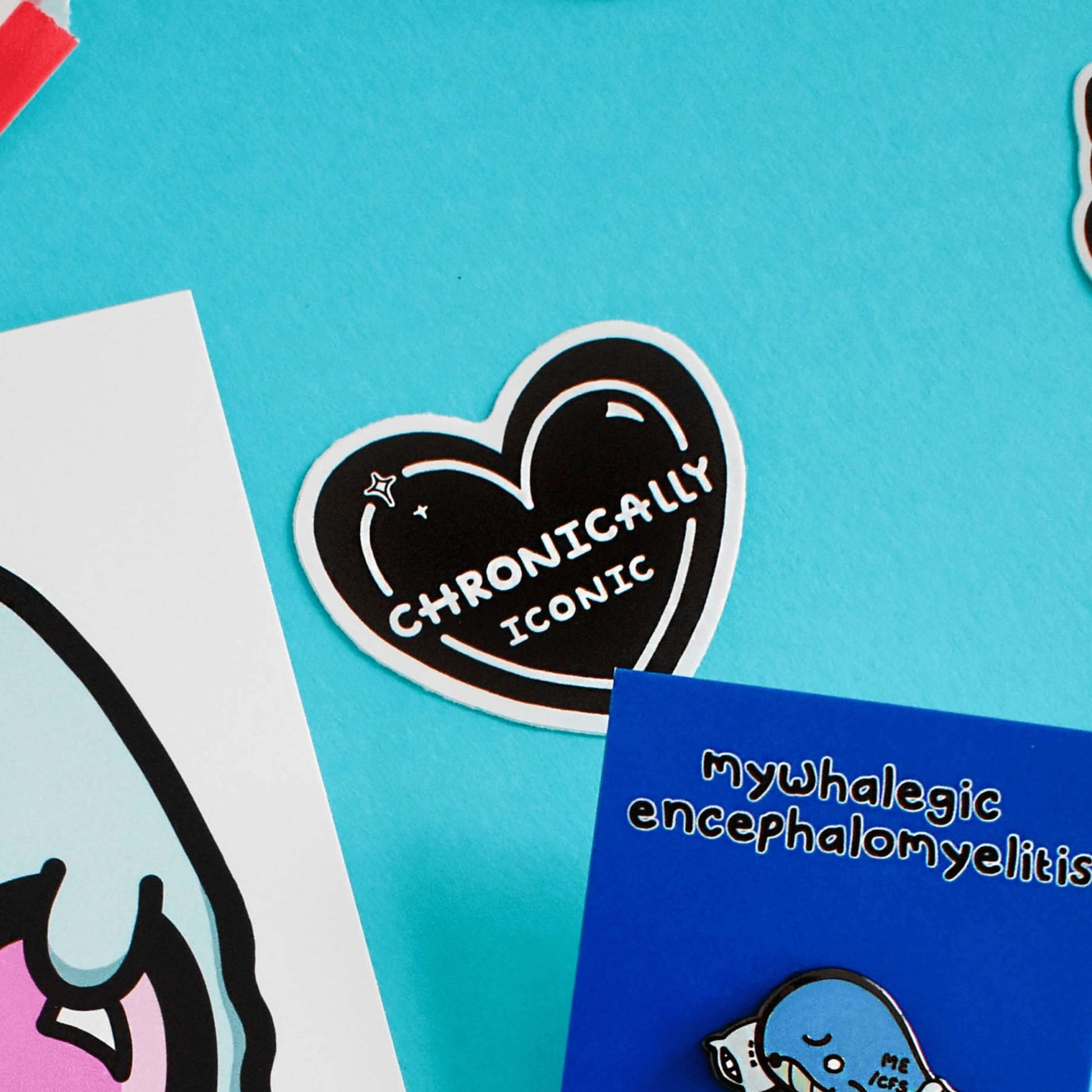 The Chronically Iconic Sticker on a blue background with other innabox products. The black heart shaped sticker has a white outline with sparkles and centre text reading 'chronically iconic'. The design is raising awareness for chronic illness and invisible illness.