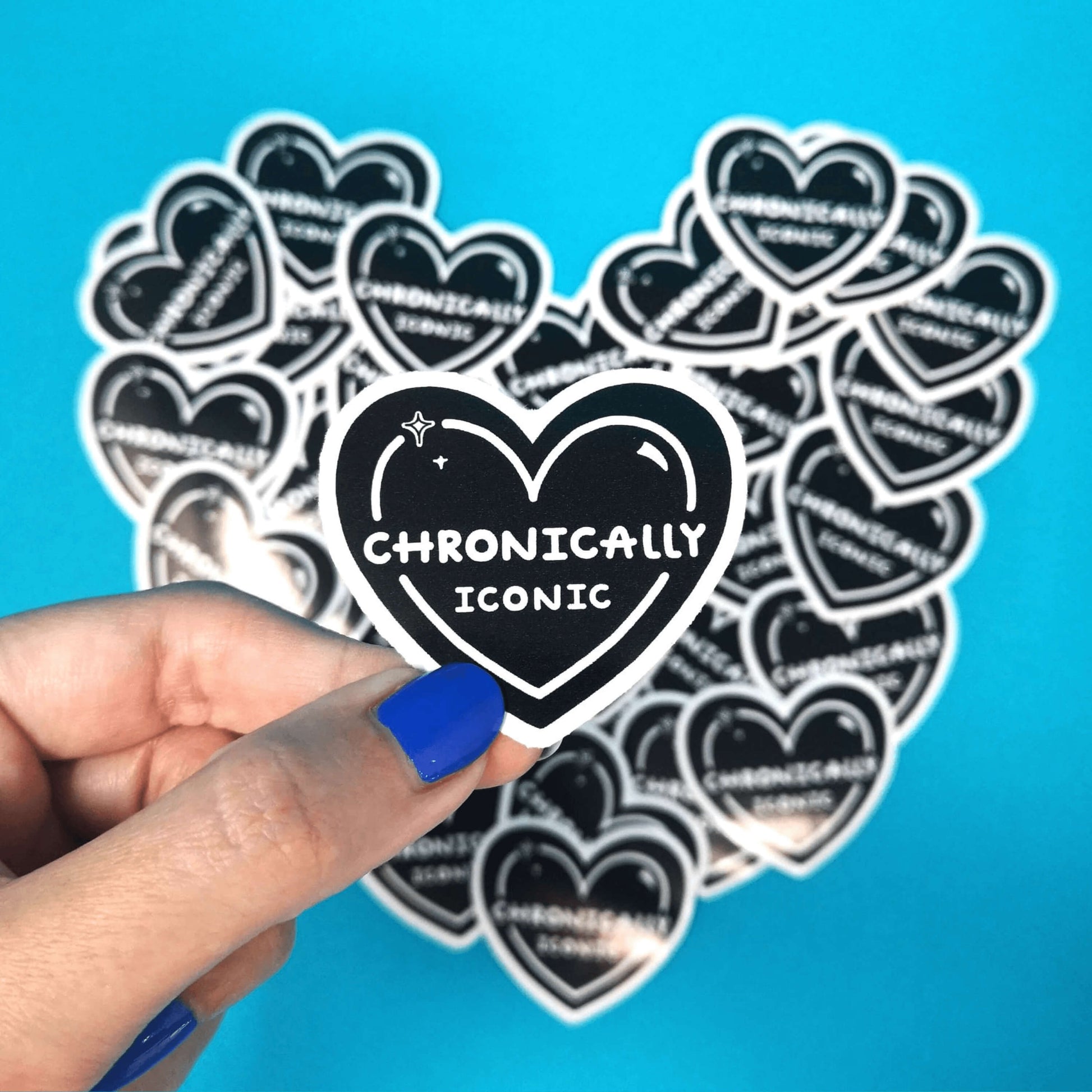 The Chronically Iconic Sticker being held over a blue background and multiple copies of the sticker pushed into the shape of a heart by a hand with blue nail varnish. The black heart shaped sticker has a white outline with sparkles and centre text reading 'chronically iconic'. The design is raising awareness for chronic illness and invisible illness.