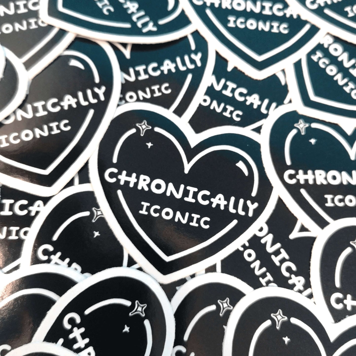 The Chronically Iconic Sticker on a pile of multiple copies of the sticker. The black heart shaped sticker has a white outline with sparkles and centre text reading 'chronically iconic'. The design is raising awareness for chronic illness and invisible illness.