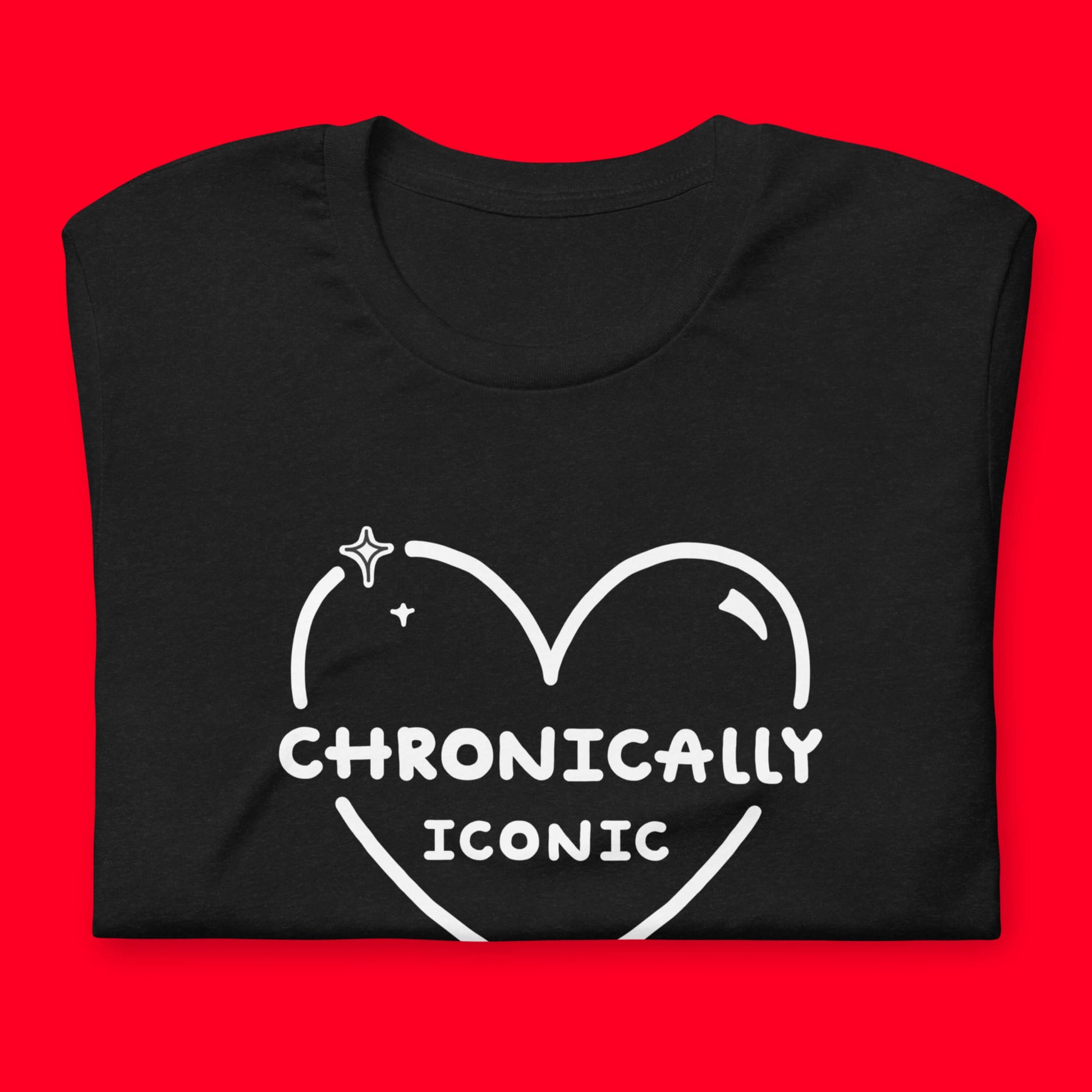 The Chronically Iconic black tee folded on a red background. The short sleeve t-shirt features a white heart outline with sparkles and centre text reading 'chronically iconic' with the innabox logo underneath. The design is raising awareness for chronic illness and invisible illness.