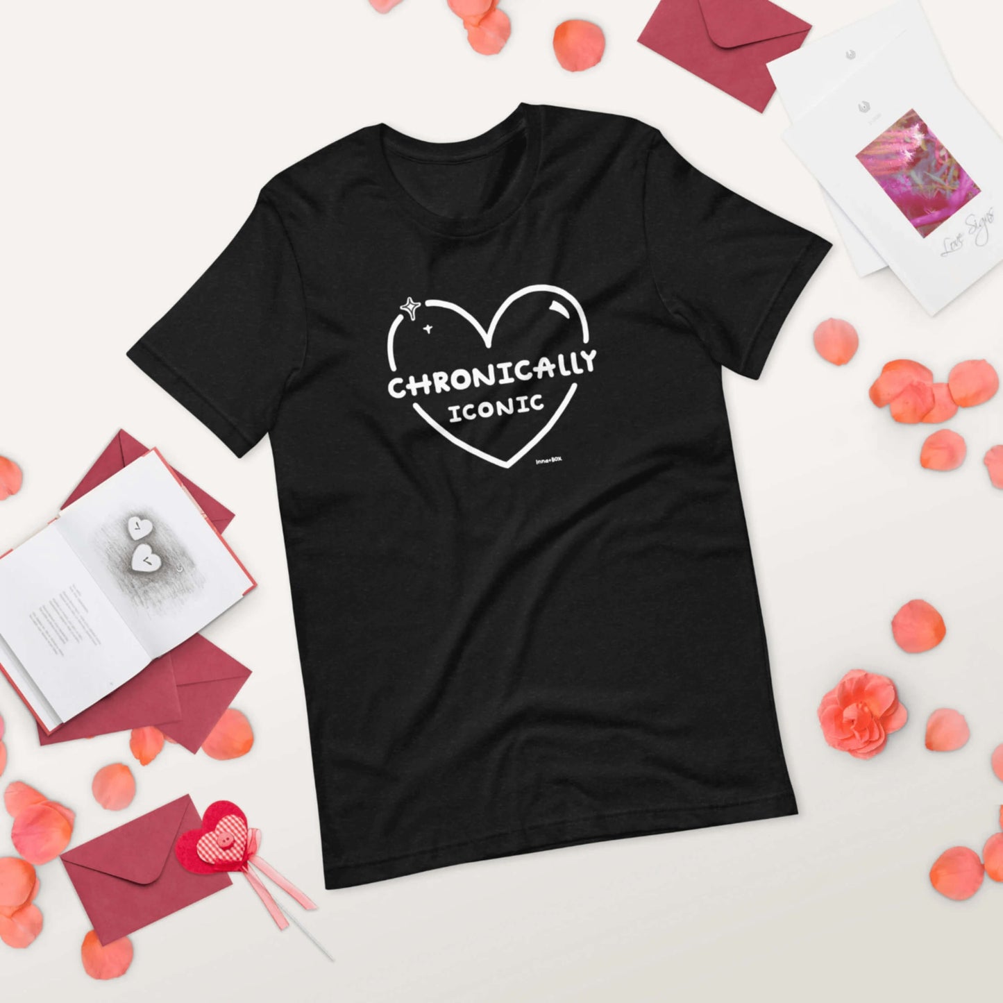 The Chronically Iconic black tee laying flat on a white background with valentines day cards, red envelopes and rose petals. The short sleeve t-shirt features a white heart outline with sparkles and centre text reading 'chronically iconic' with the innabox logo underneath. The design is raising awareness for chronic illness and invisible illness.