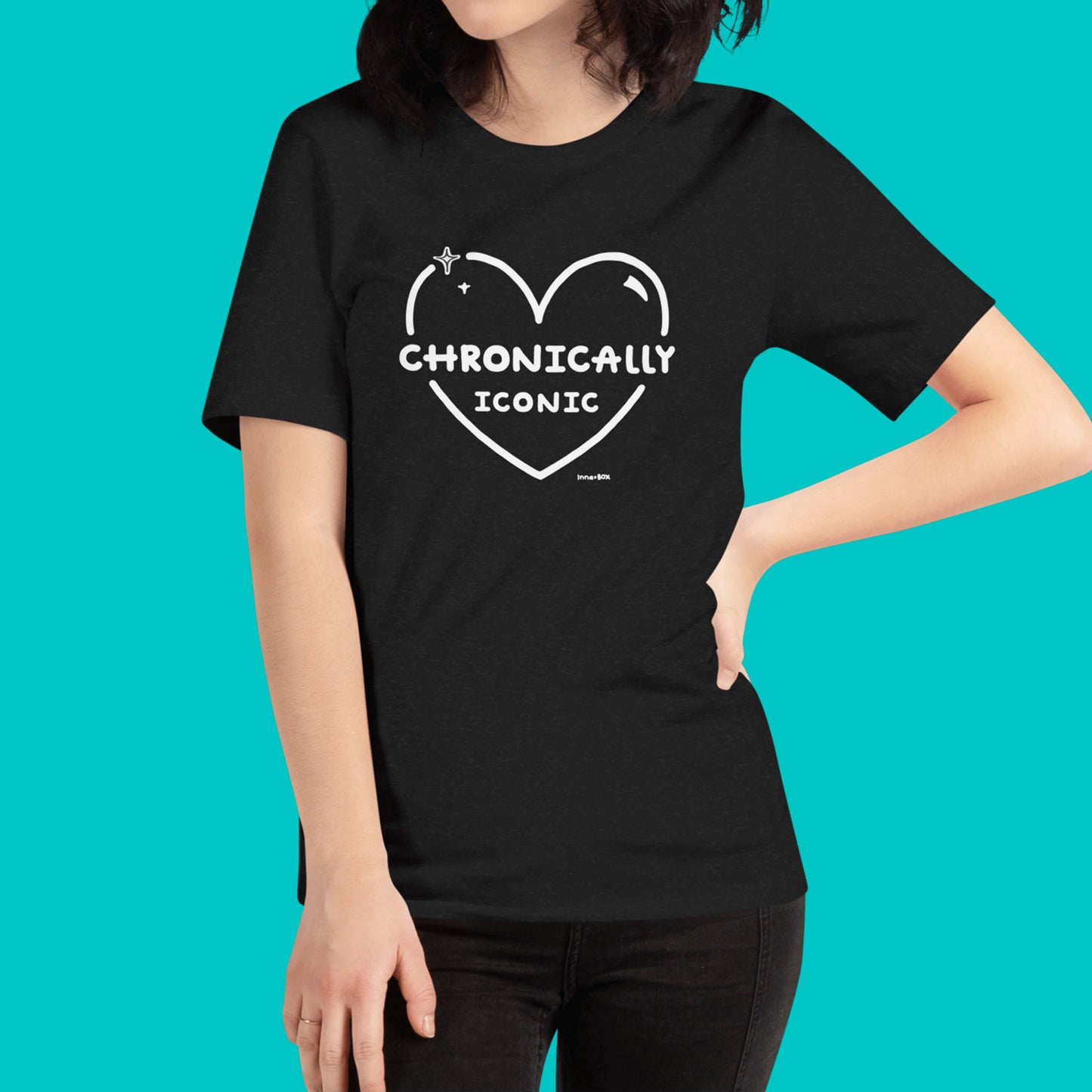 The Chronically Iconic black tee worn by a model with brown short hair on a blue background. The short sleeve t-shirt features a white heart outline with sparkles and centre text reading 'chronically iconic' with the innabox logo underneath. The design is raising awareness for chronic illness and invisible illness.