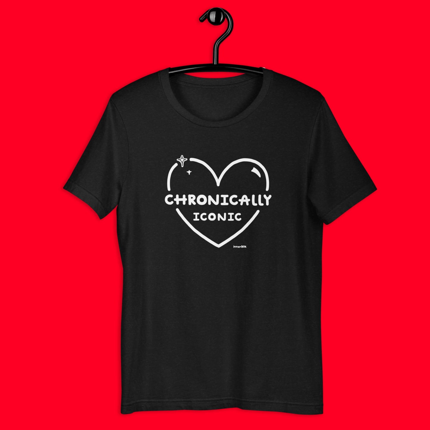 The Chronically Iconic black tee hanging up on a black hanger over a red background. The short sleeve t-shirt features a white heart outline with sparkles and centre text reading 'chronically iconic' with the innabox logo underneath. The design is raising awareness for chronic illness and invisible illness.