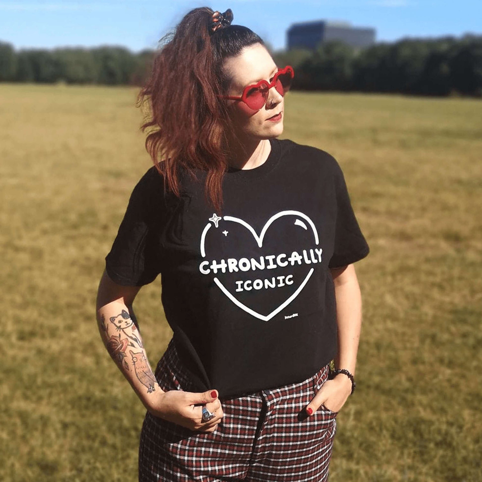 The Chronically Iconic black tee modelled by Nikky, with long brown hair in a ponytail and tattoos. She is facing forward looking off to the right pulling on the tucked in tshirt, she is wearing a black scrunchie, red heart sunglasses and red tartan high waist trousers. The short sleeve t-shirt features a white heart outline with sparkles and centre text reading 'chronically iconic' with the innabox logo underneath. The design is raising awareness for chronic illness and invisible illness.