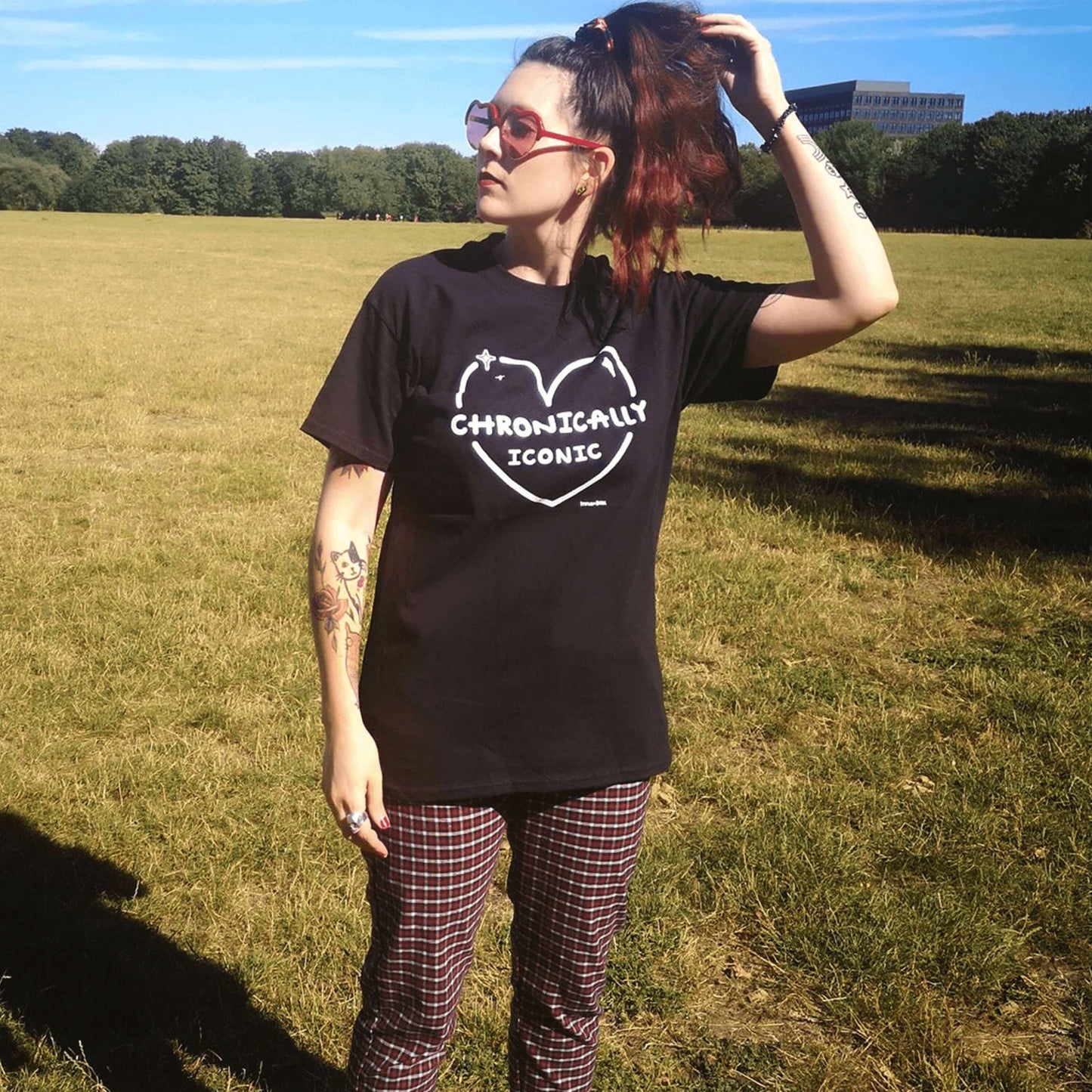 The Chronically Iconic black tee modelled by Nikky, with long brown hair in a ponytail and tattoos. She is facing forward looking off to the left whilst brushing her hand through her ponytail, she is wearing a black scrunchie, red heart sunglasses and red tartan high waist trousers. The short sleeve t-shirt features a white heart outline with sparkles and centre text reading 'chronically iconic' with the innabox logo underneath. The design is raising awareness for chronic illness and invisible illness.