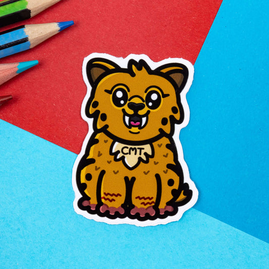 The Charcot Sabre-Tooth Tiger Sticker - Charcot-Marie-Tooth Disease CMT on a blue and red background with colouring pencils. A brown smiling sabre-tooth sticker with the initials 'CMT' in black across its chest. The hand drawn design is raising awareness for Charcot-Marie-Tooth Disease CMT.